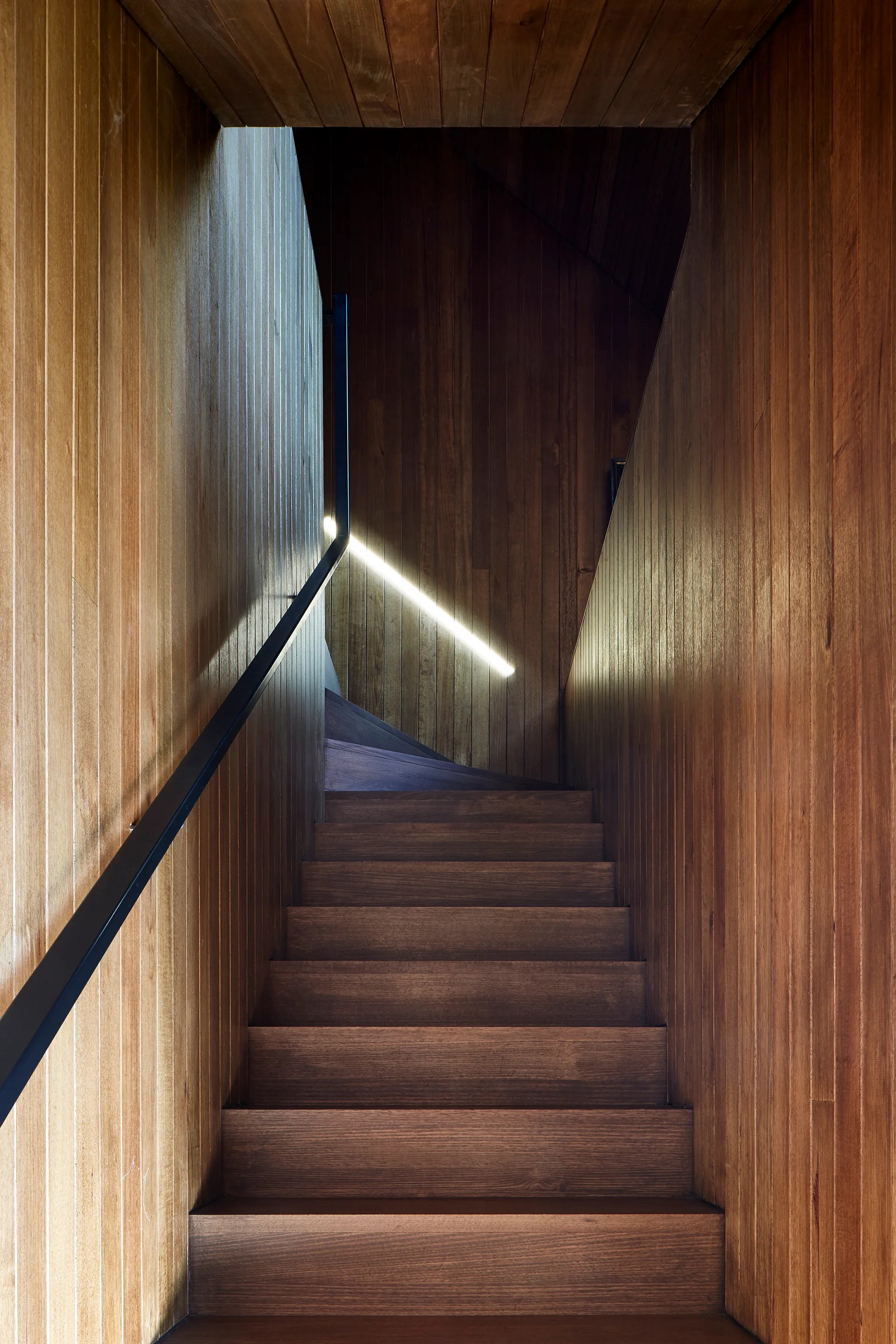 A minimalist wooden staircase features vertical wood slat wall cladding, a simple black metal handrail, and a single diagonal glowing linear light strip.