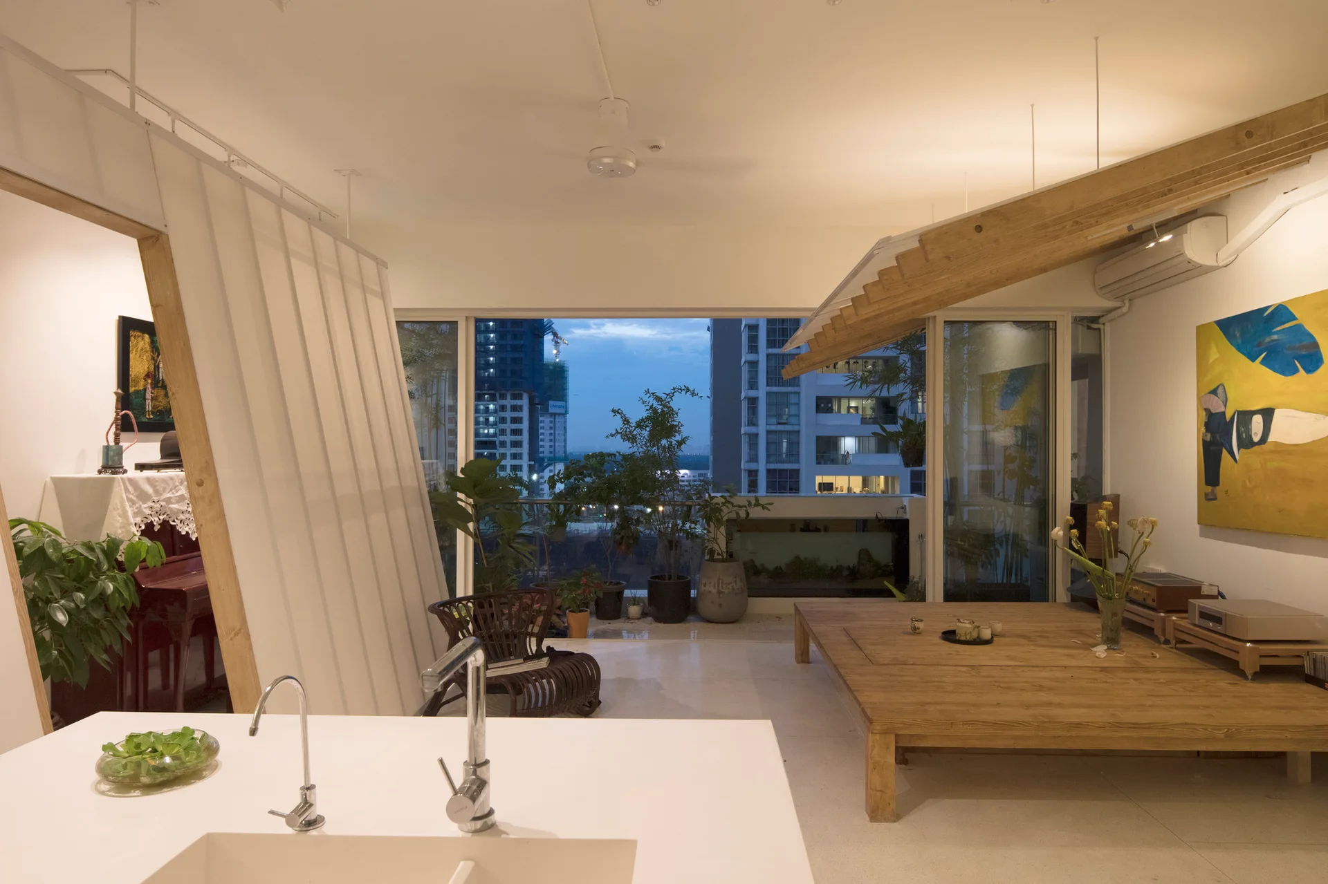 An open apartment living space with a low wooden platform, a white kitchen counter, a slatted partition, and large glass doors showing a city skyline.