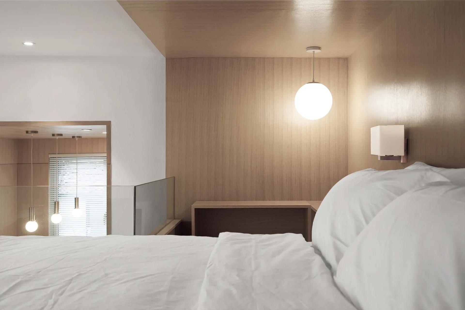 A lofted bedroom features crisp white bedding beneath a warm wood-paneled wall and ceiling accented by a glowing globe pendant light.