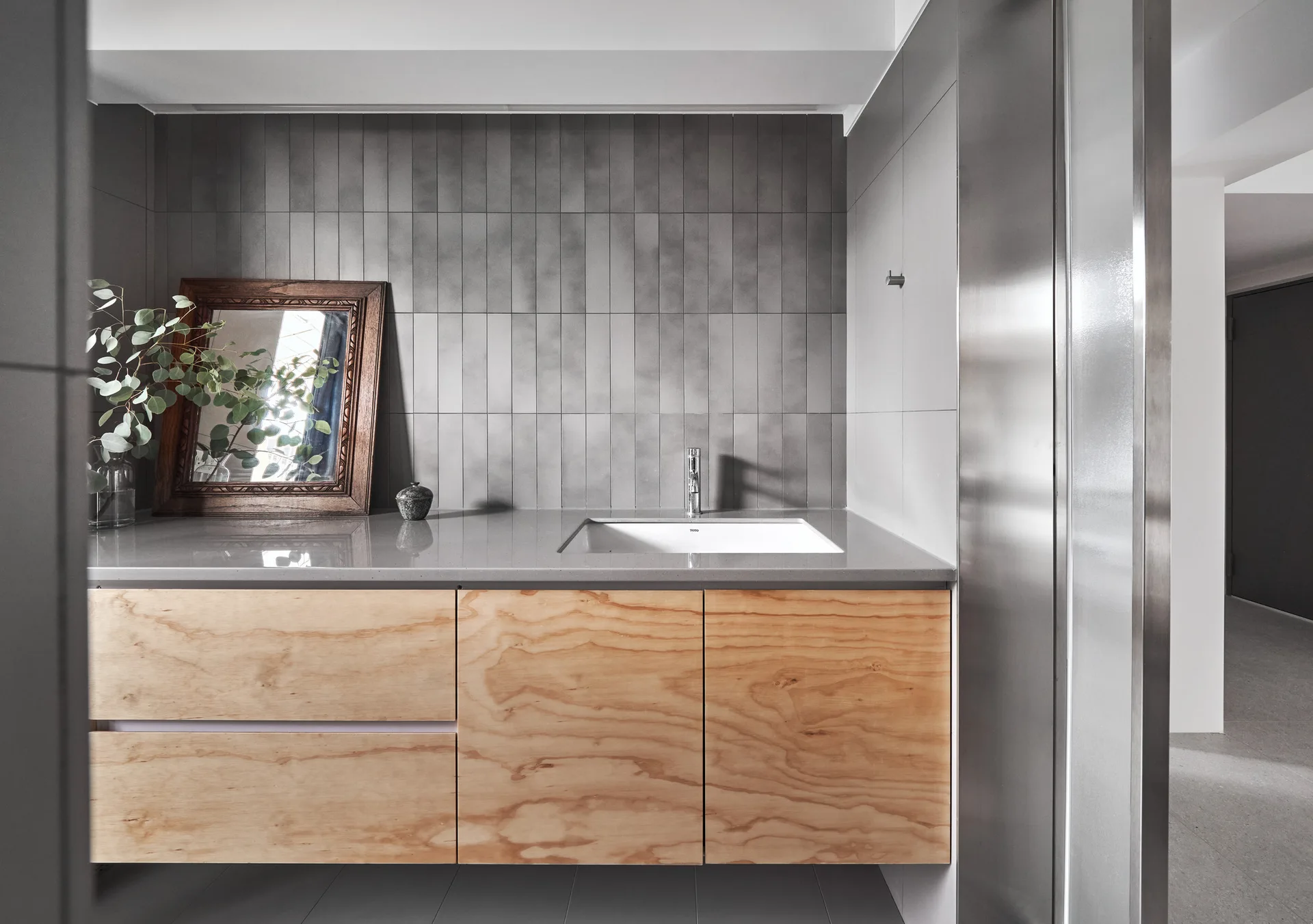 A modern bathroom vanity features light wood cabinet doors, a gray countertop with an undermount sink, and a vertically tiled gray accent wall.