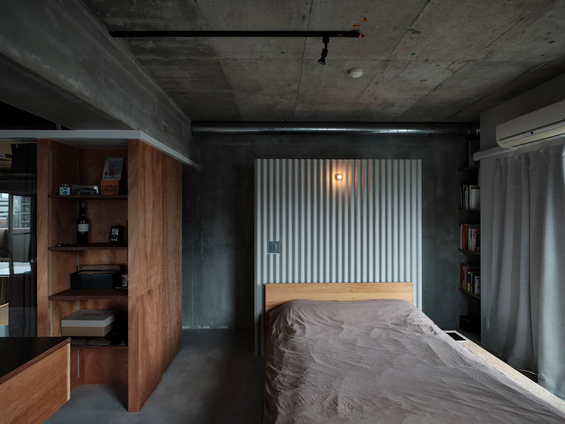 A minimalist bedroom features a wooden bed set against a corrugated metal panel on raw concrete walls, alongside a wooden storage cabinet.