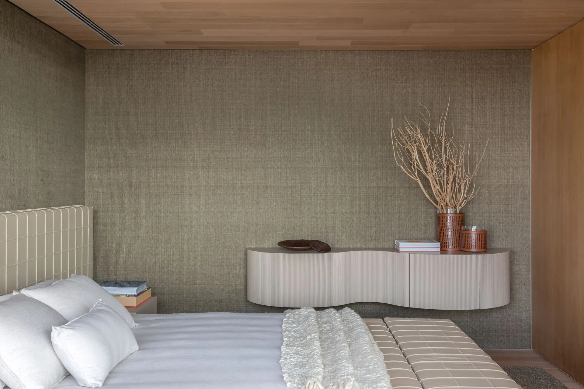 A minimalist bedroom featuring a textured fabric wall covering, a curved floating console with dried branches, and an upholstered bed.