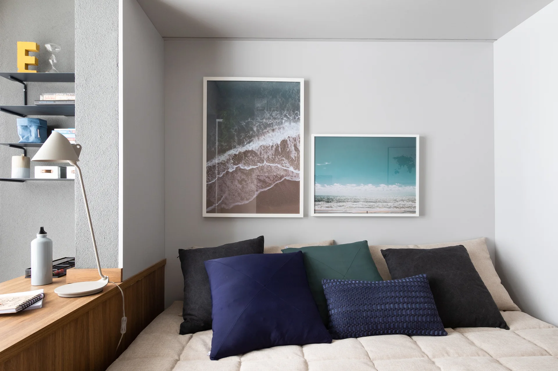 A grey bedroom alcove features a beige daybed with dark blue and green pillows under two framed ocean photos, next to a wood desk with a white lamp.