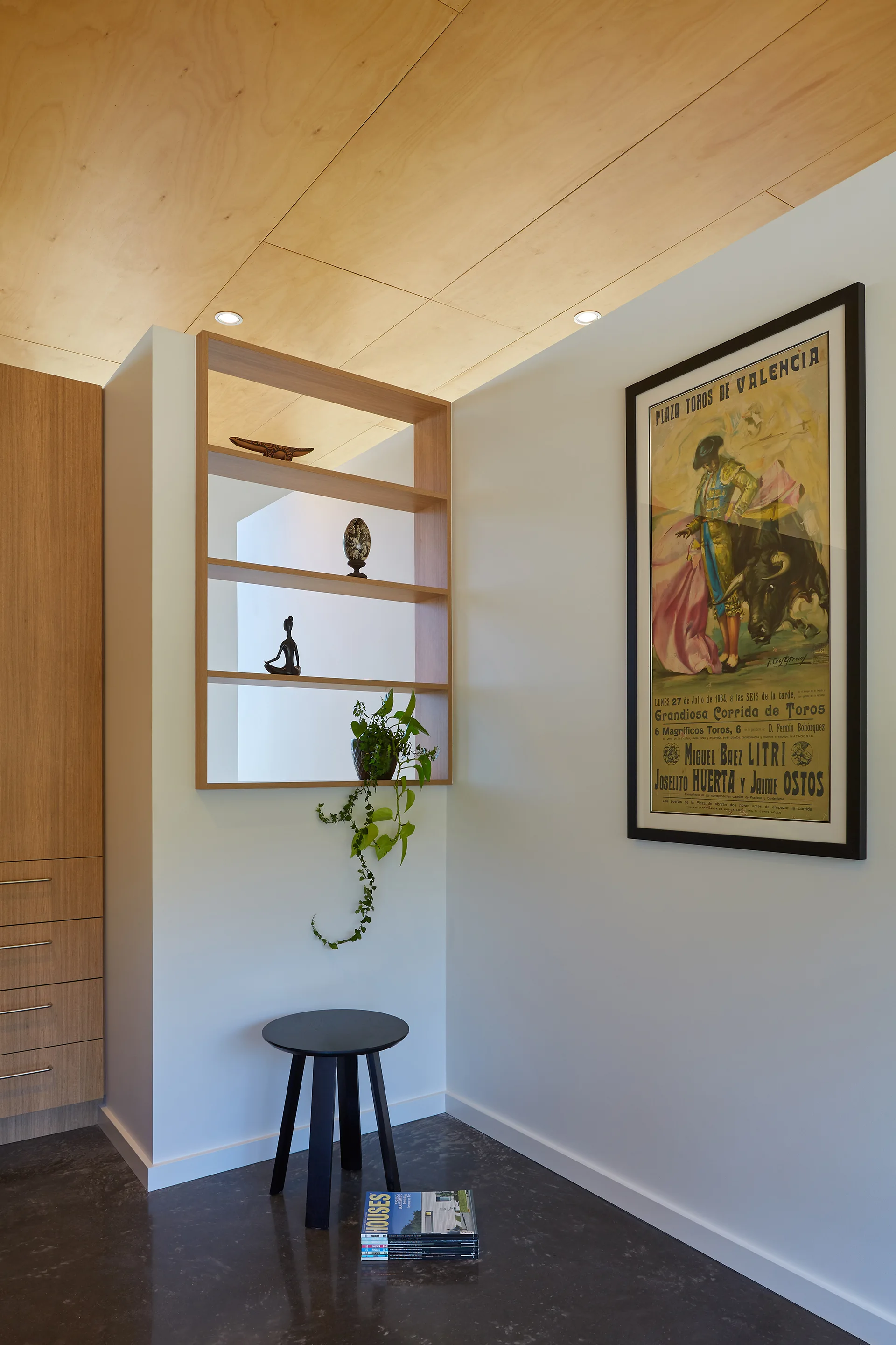 A modern interior corner featuring a wood panel ceiling, recessed wood shelves, a black stool, and a framed vintage poster on a white wall.