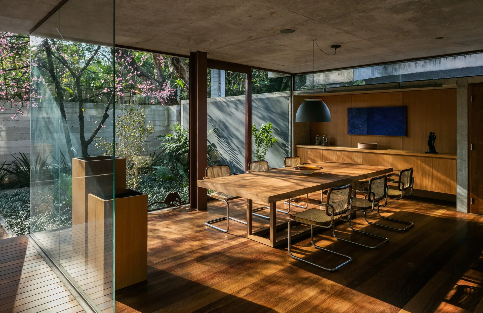 A modern dining room features a wooden table and cane chairs set against floor-to-ceiling glass walls overlooking a garden.