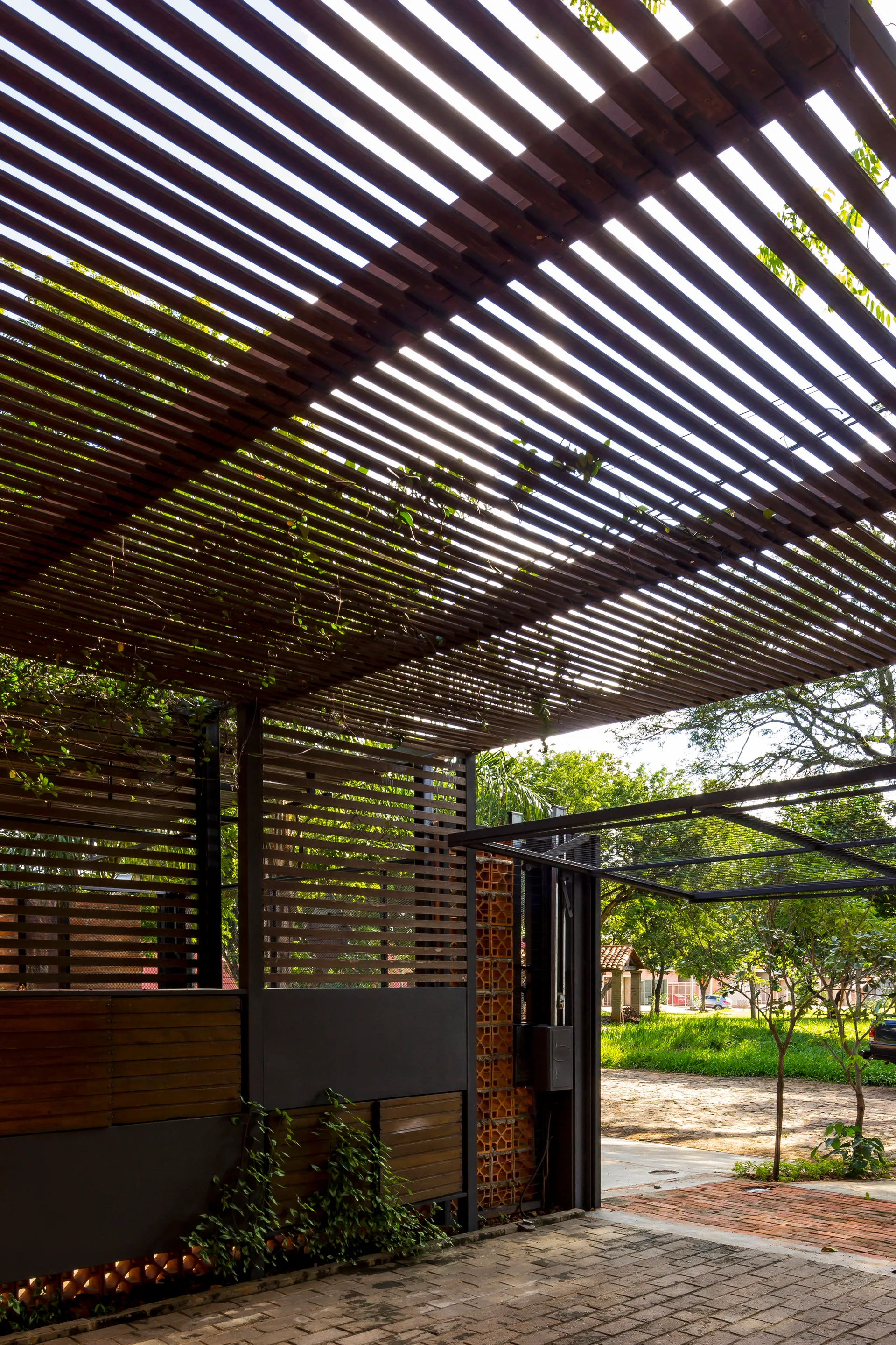 A paved outdoor driveway features a large dark wooden slatted pergola roof overhead next to a terracotta lattice screen wall and a metal gate.