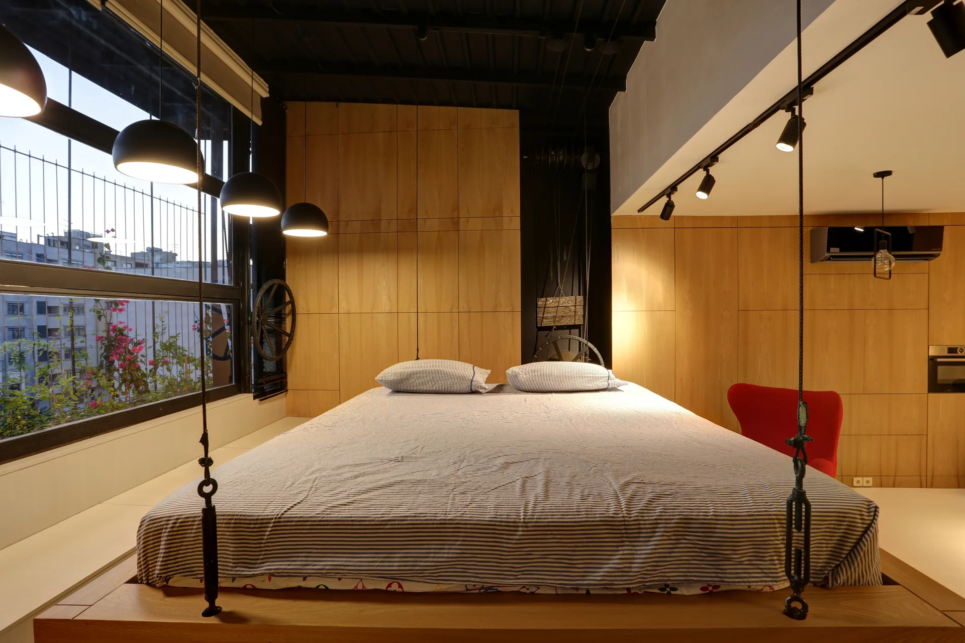 A low wood platform bed with striped linens sits against wood-paneled walls in a bedroom with a dark exposed industrial ceiling.