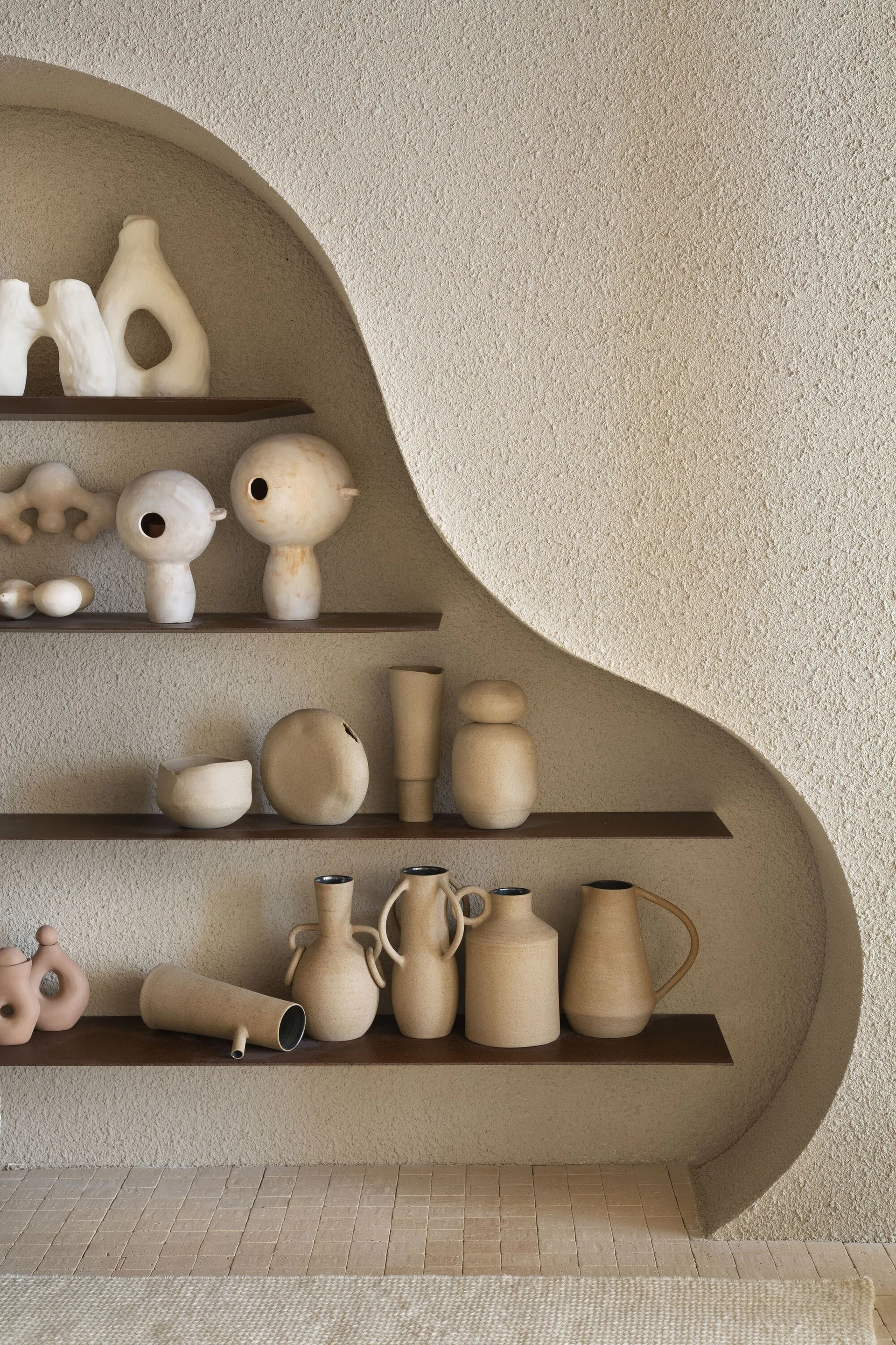 A collection of earth-toned ceramic vases and abstract sculptures displayed on dark wood shelves recessed within a textured plaster archway.