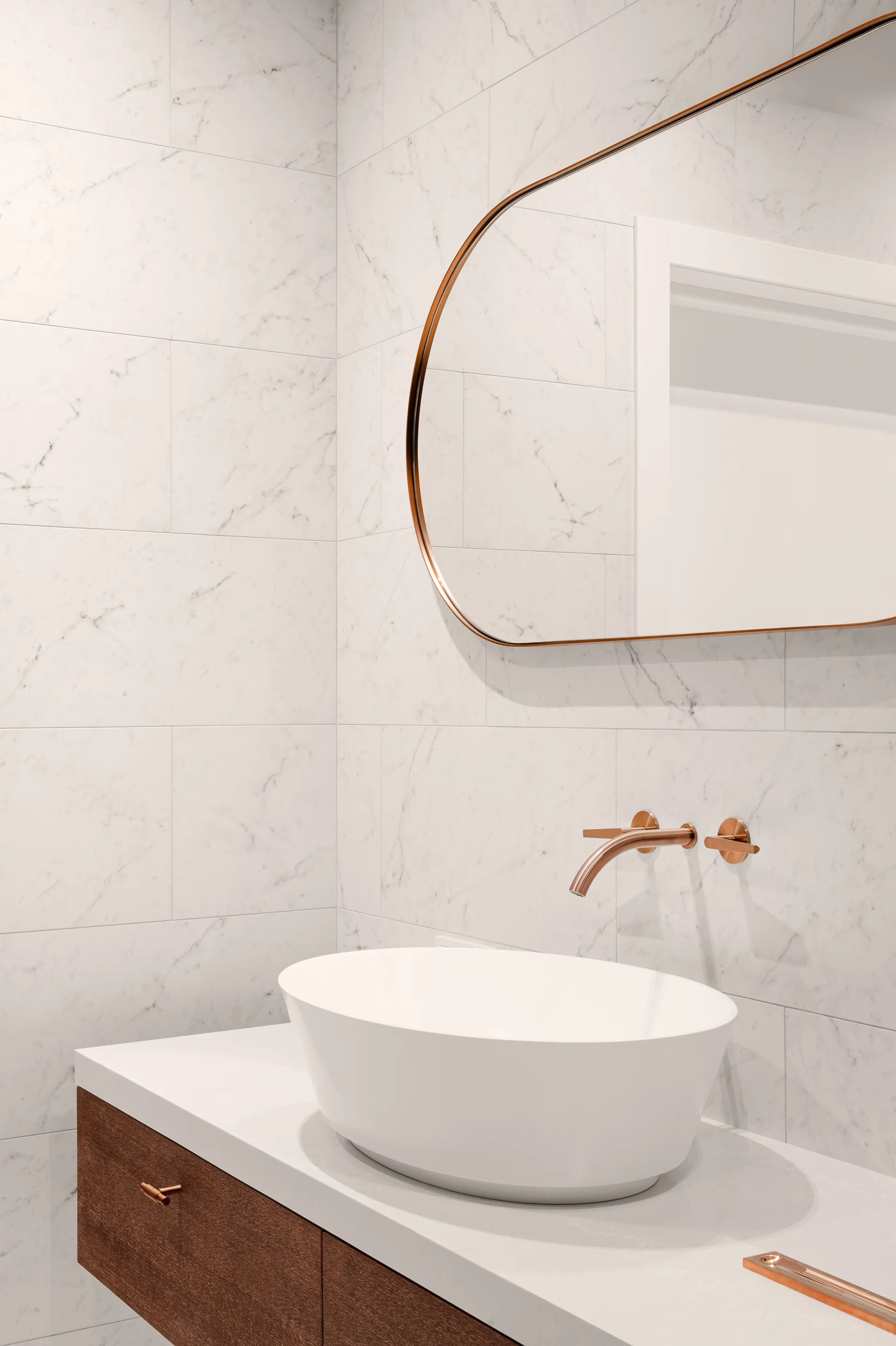 A close-up shot of a modern bathroom vanity featuring a white vessel sink, copper wall-mounted faucet, pill-shaped mirror, and veined marble tile walls.