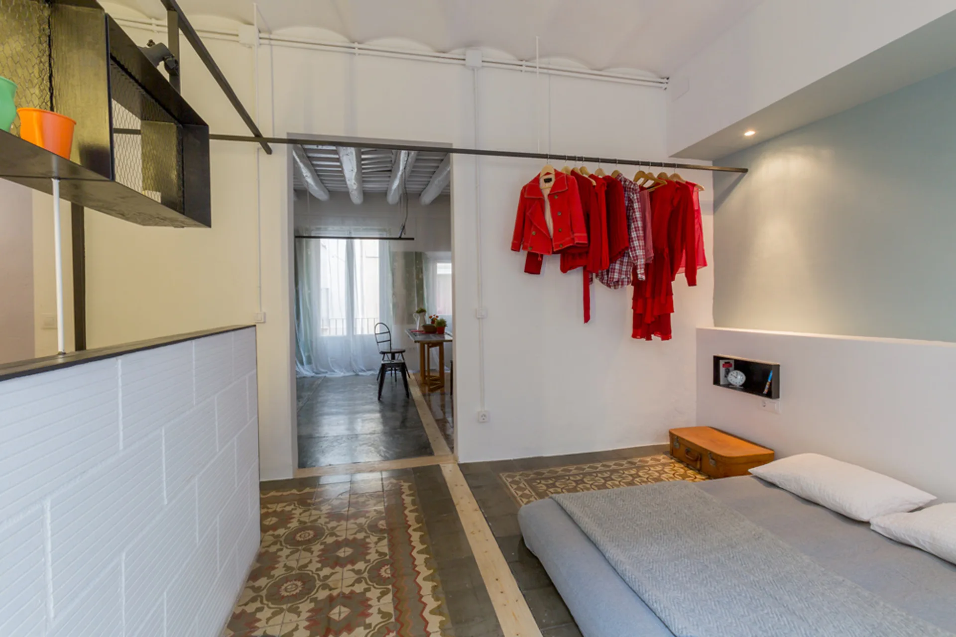 A minimalist bedroom features a low grey bed, patterned geometric floor tiles, and a black wall-mounted metal rail displaying red clothes.