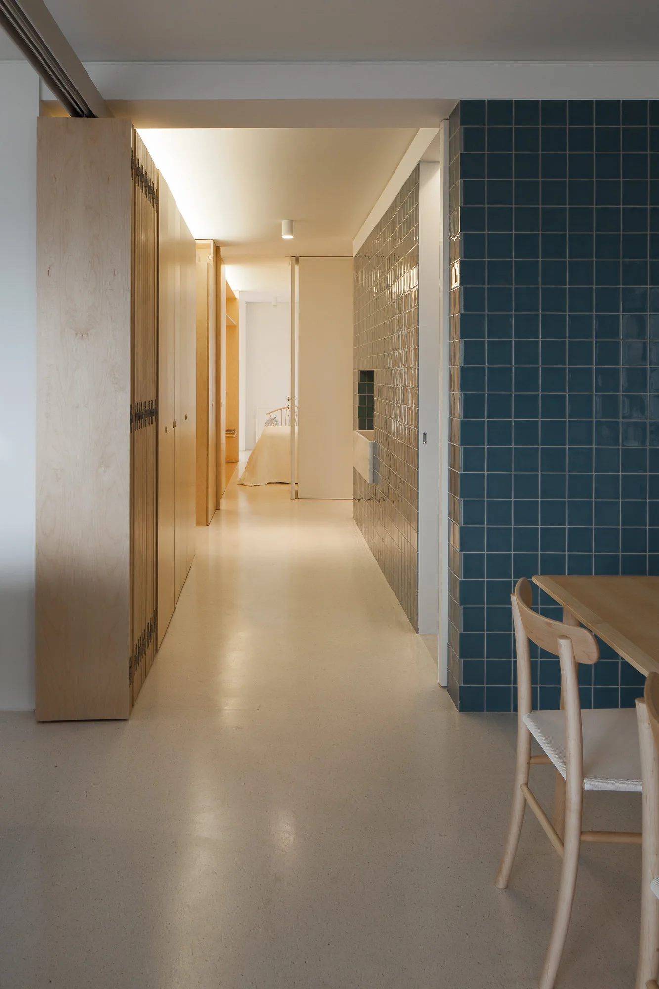 A minimalist hallway features tall light wood cabinetry on the left and a glossy blue tiled wall partition next to a dining table on the right.