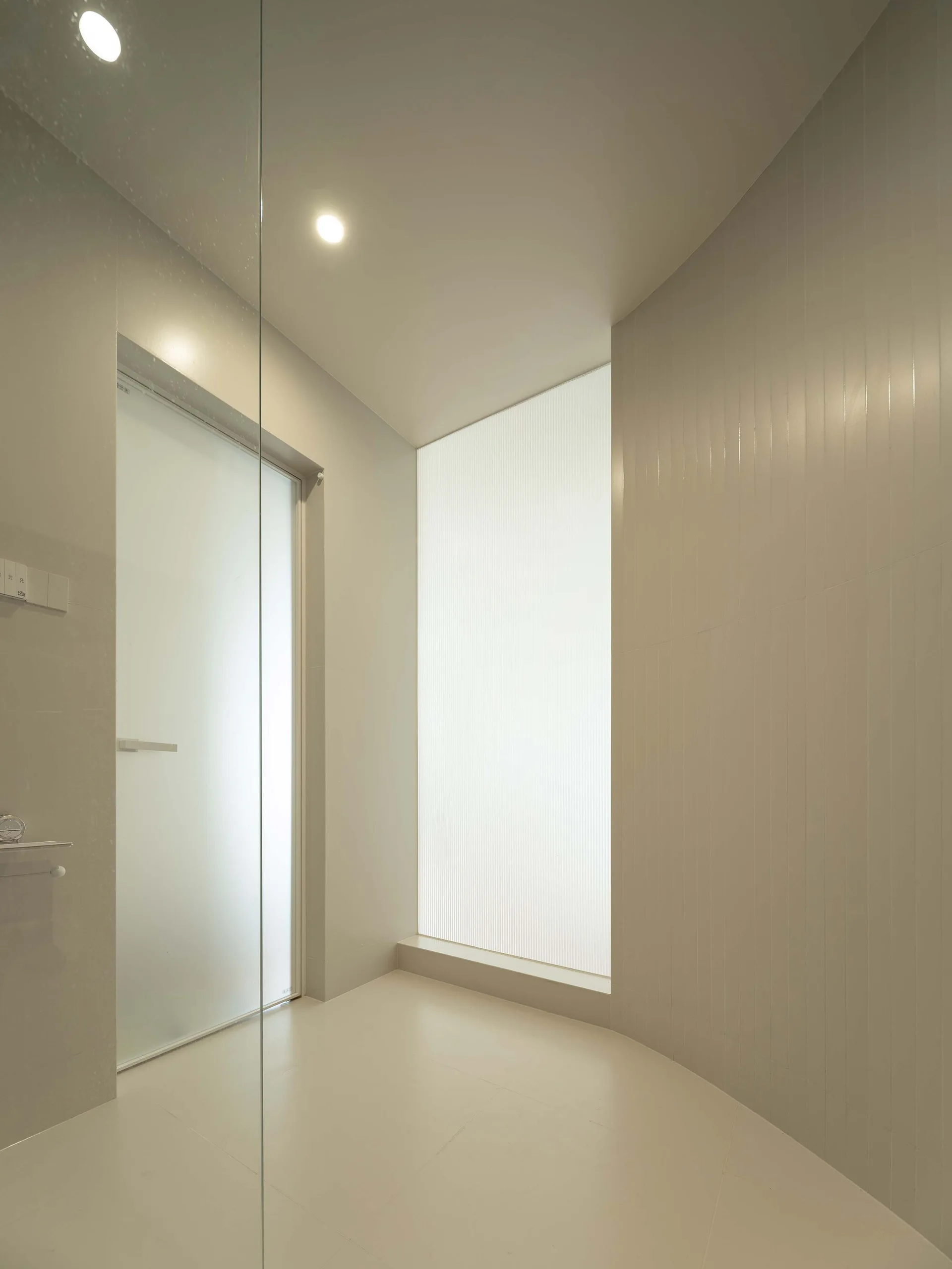 A minimalist beige shower area featuring vertical tiled walls, a fluted glass window panel, a frosted glass door, and recessed ceiling lights.