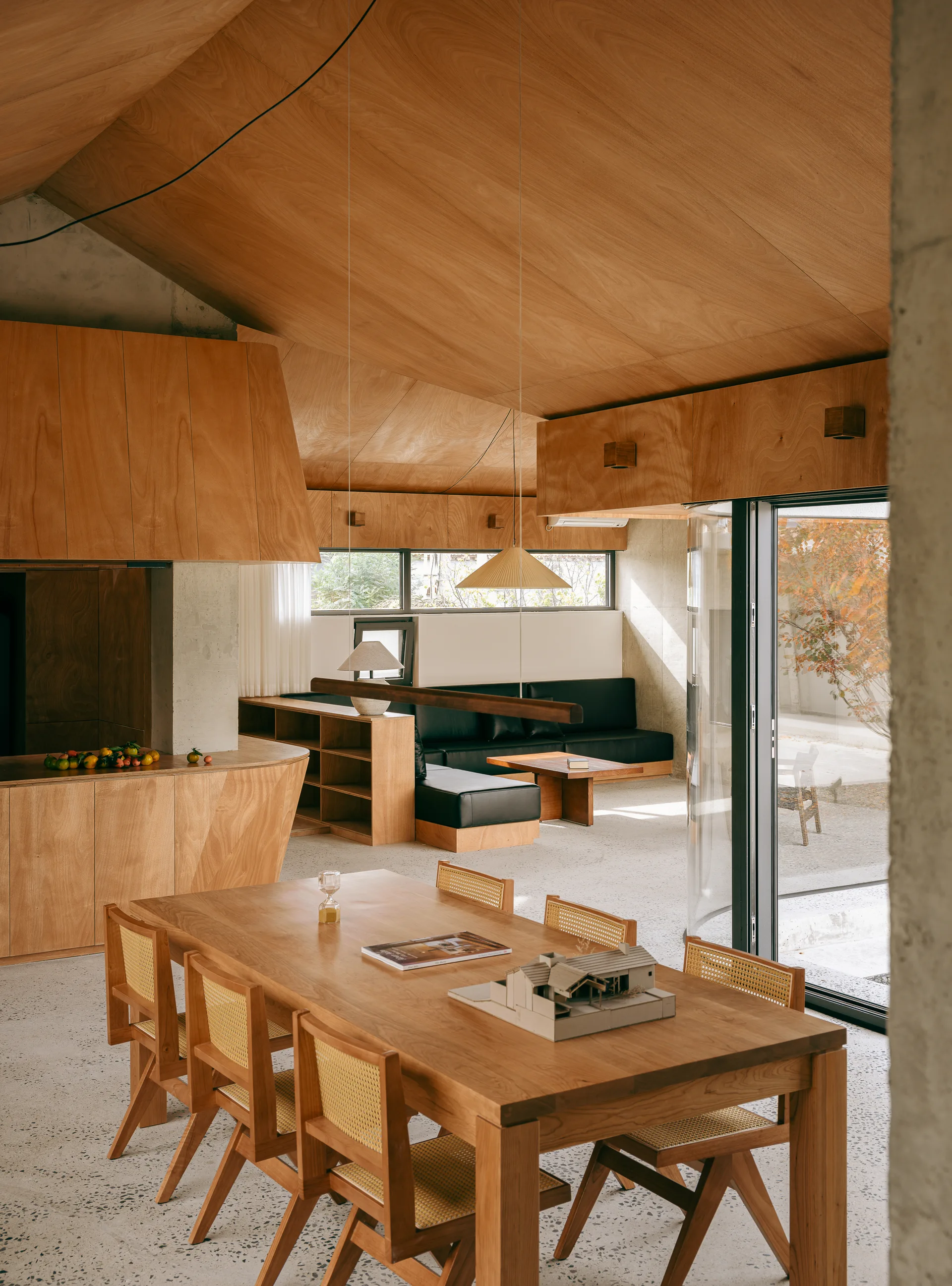 A minimalist dining room showcases a wooden table with cane chairs set beneath a sloped plywood ceiling and concrete columns.