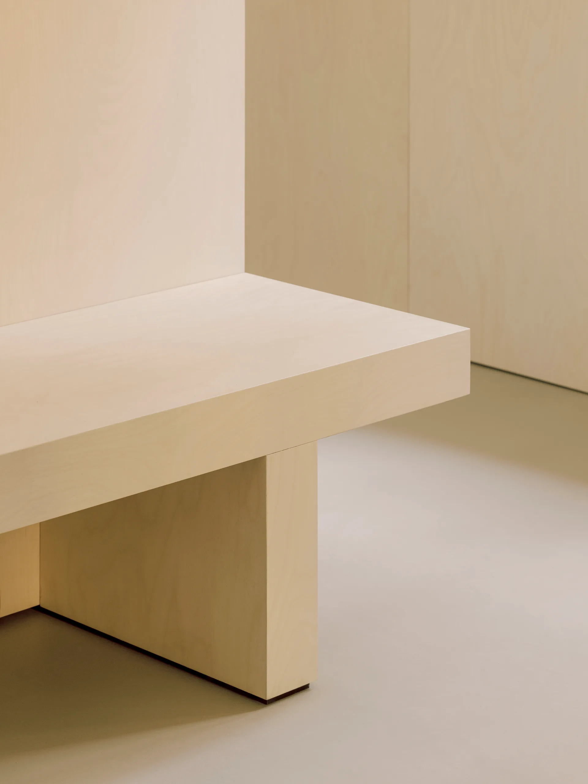 A minimalist light wood bench stands against matching blonde wood wall panels on a smooth matte beige floor.
