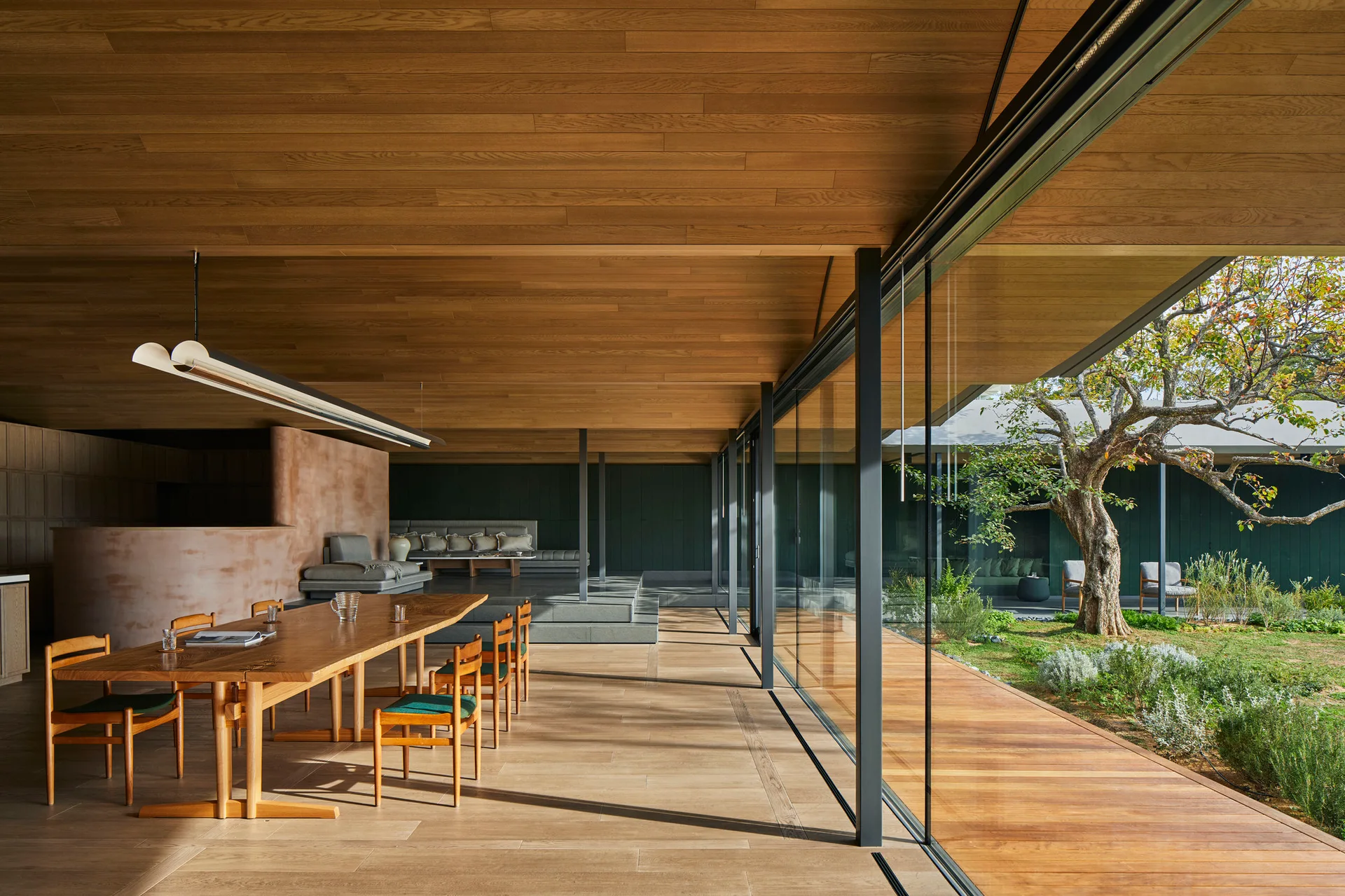 A modern dining room features a wooden table and chairs beneath a wood-plank ceiling, with floor-to-ceiling glass walls looking out to a garden.