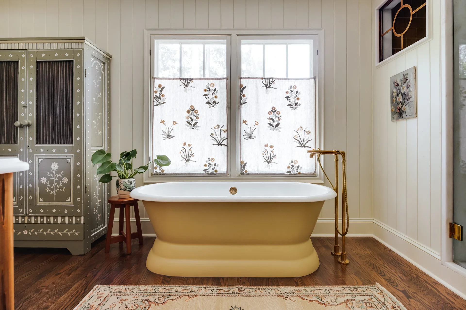 A cottage bathroom featuring a freestanding yellow pedestal bathtub under a window with floral cafe curtains, next to a tall painted cabinet.