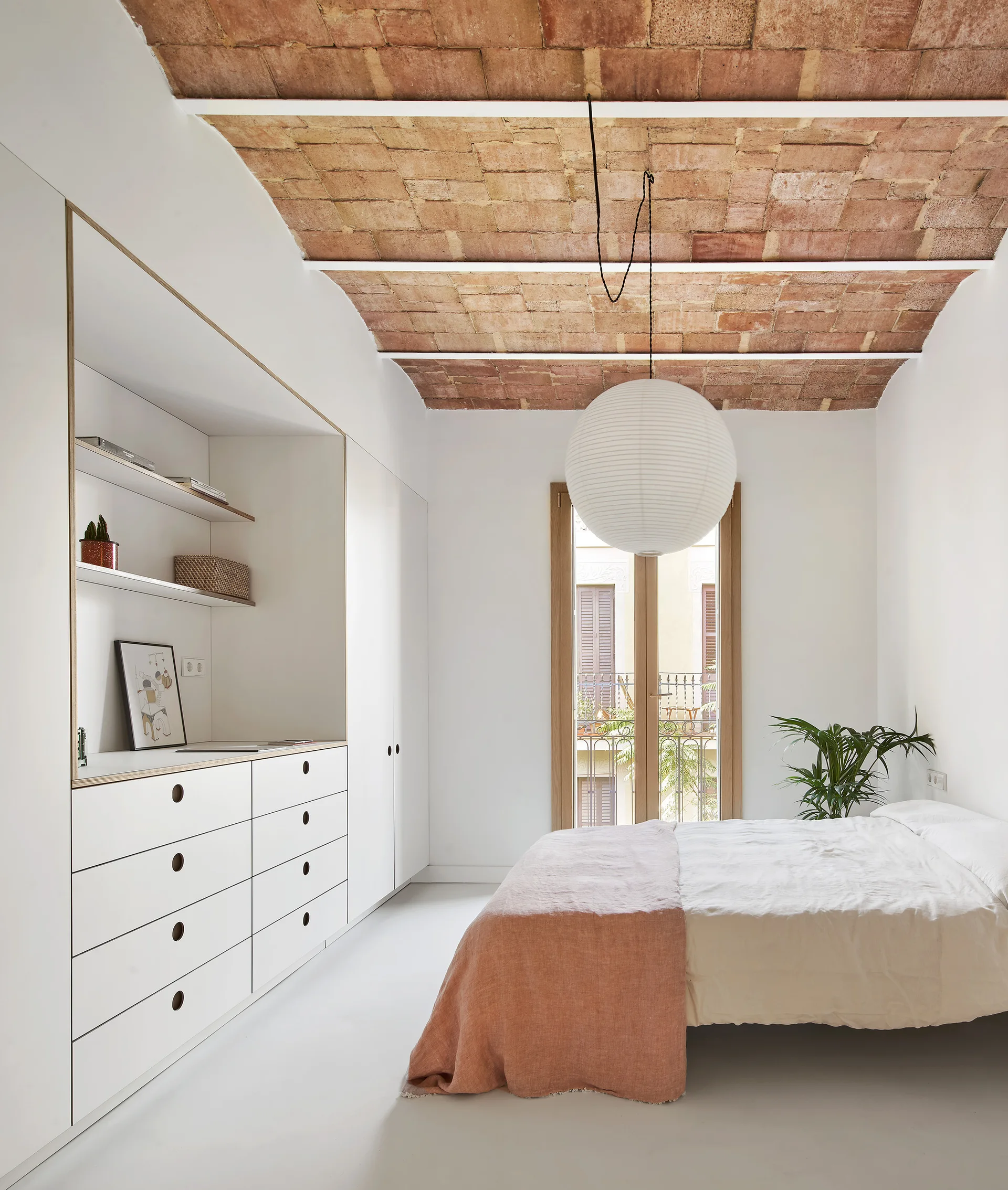 A minimalist bedroom featuring an exposed brick vaulted ceiling, built-in white cabinets, and a bed beneath a large paper pendant light.