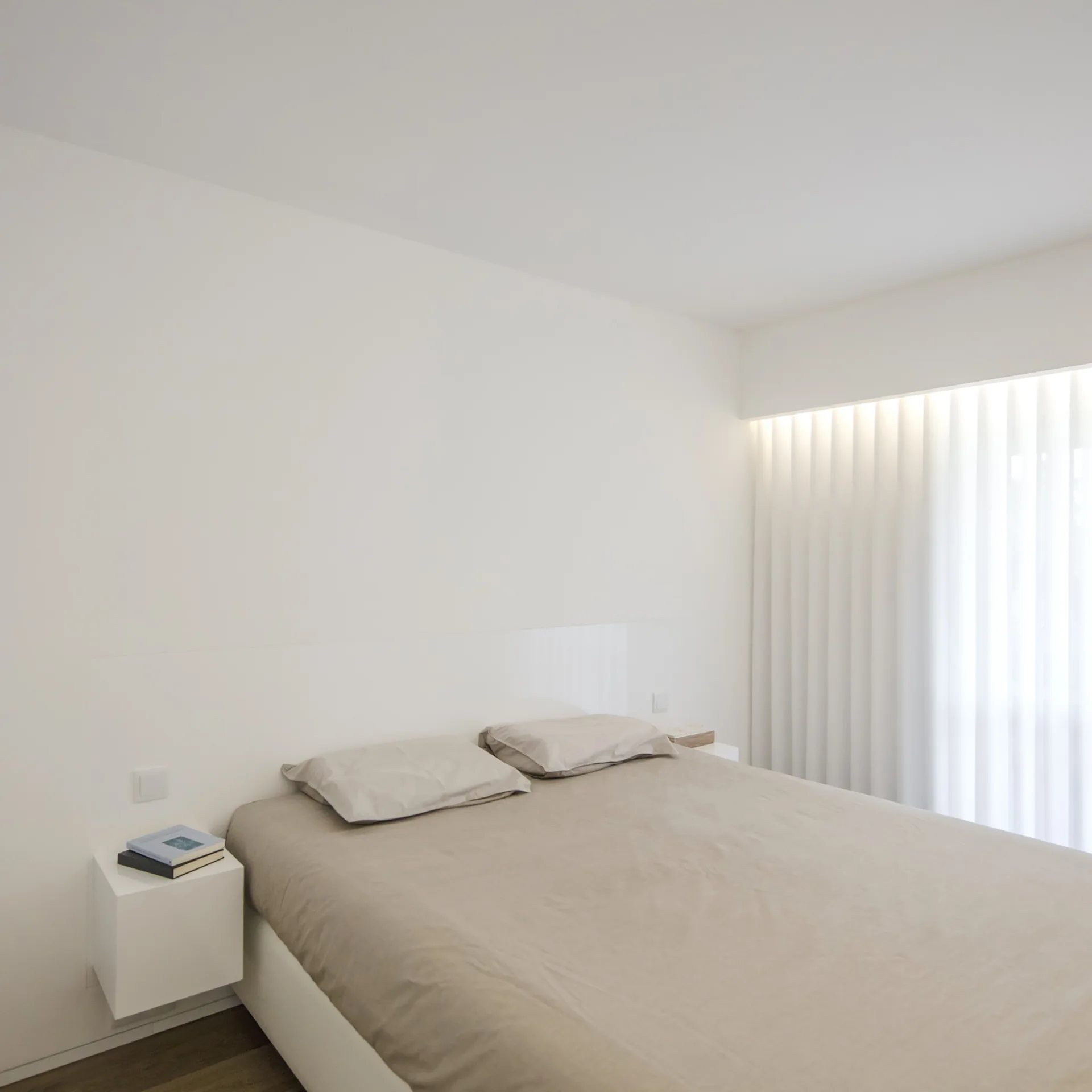 A minimalist white bedroom features a bed with beige linens next to a floating white nightstand, with sheer white curtains illuminated by warm cove lighting.