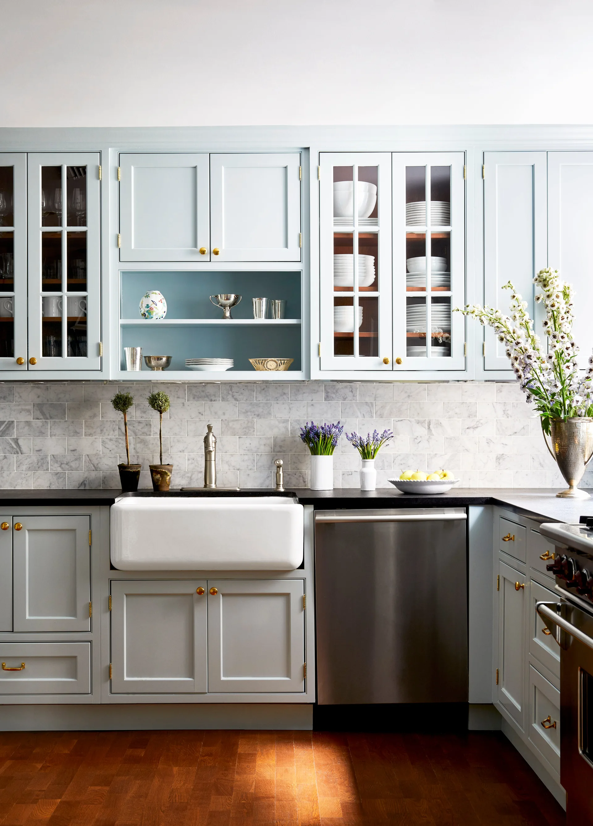 A kitchen features light blue cabinets, a white farmhouse sink, a dark countertop, and a marble subway tile backsplash.