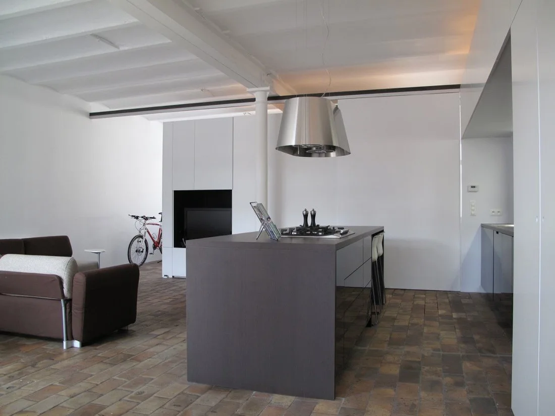 A modern open-plan kitchen features a dark gray island, white painted ceiling beams, a brown sofa, and rustic terracotta tile flooring.