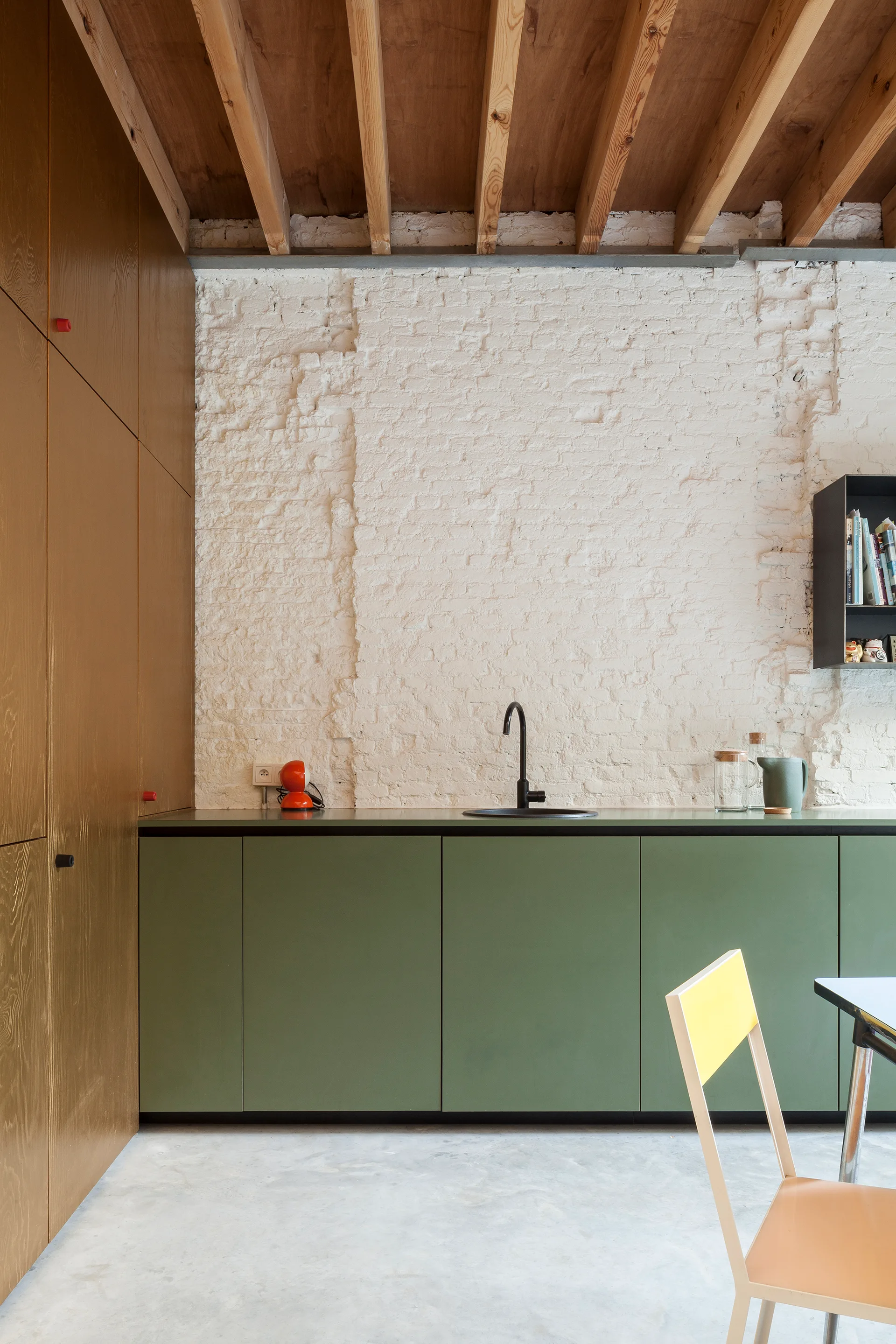 A modern kitchen featuring matte green base cabinets, a white painted brick wall, a black gooseneck faucet, and exposed natural wood ceiling beams.