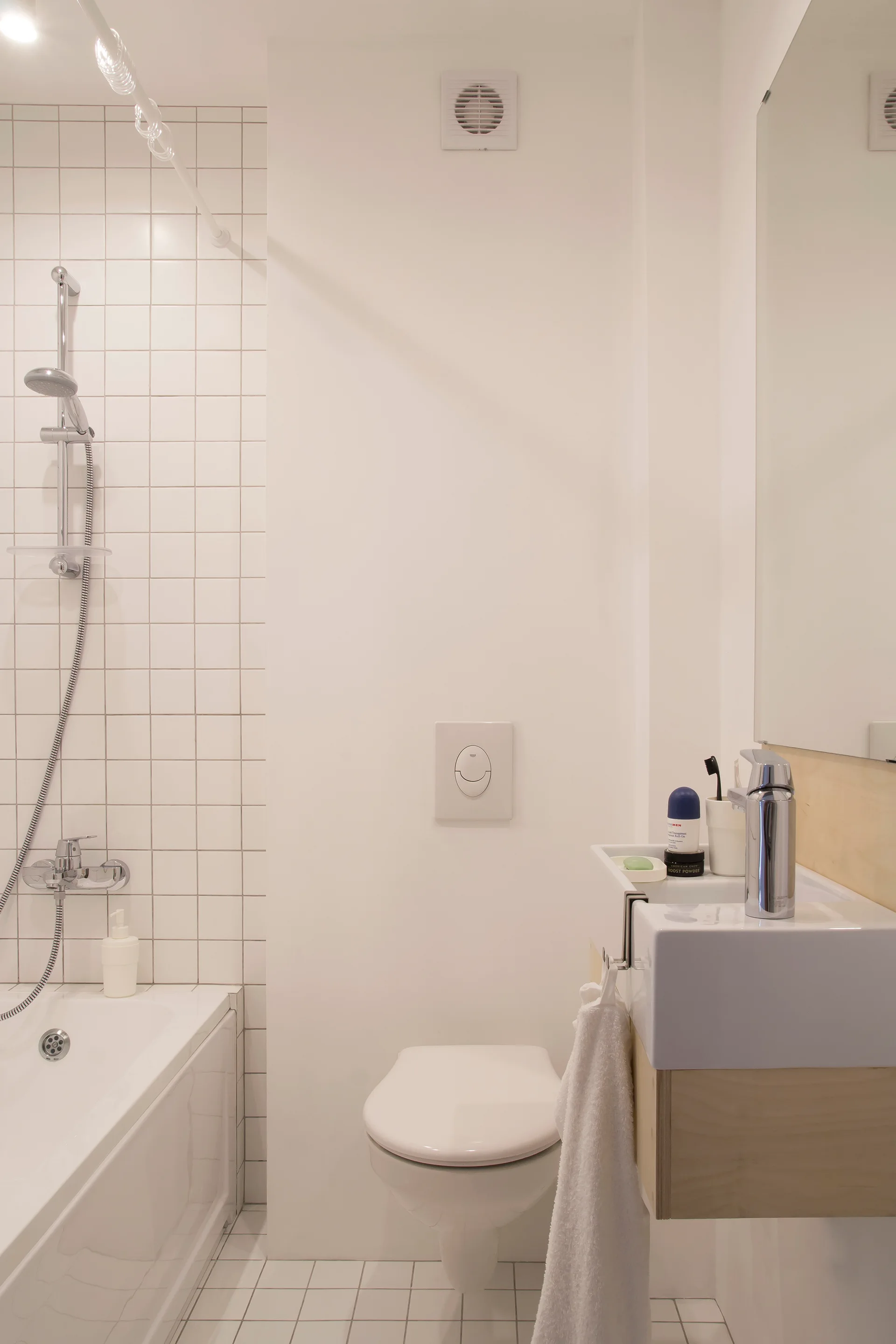 A small white bathroom featuring a wall-mounted toilet, a compact sink with light wood trim, and square grid tiling in the shower.