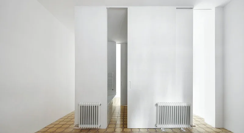 A minimalist white hallway features large sliding door panels, column radiators, and a beige grid-patterned tile floor.