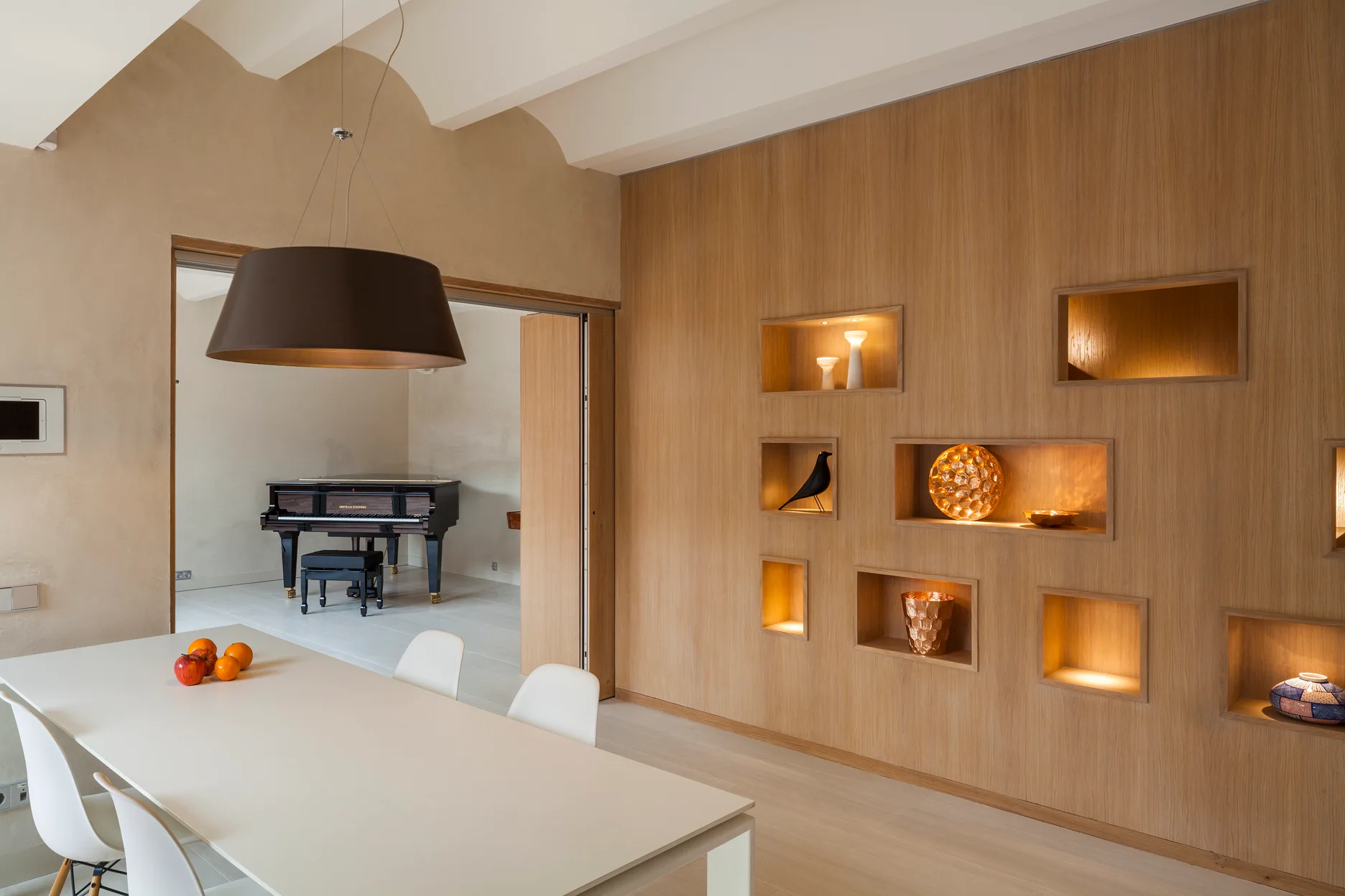 A white modern dining table set sits beside a wood-paneled wall with warm backlit recessed display niches, showing a grand piano in the background.