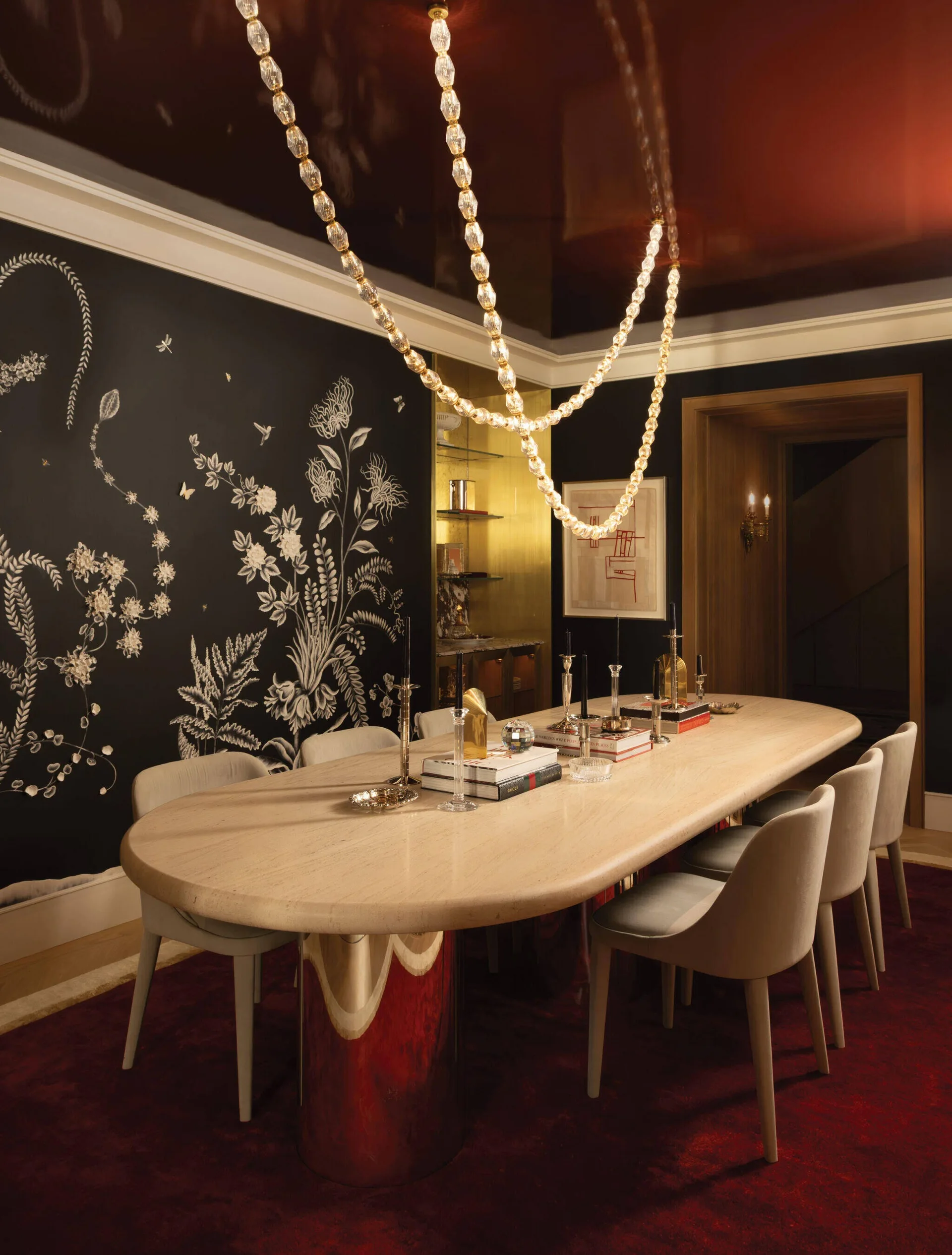A dining room features a long oval stone table, upholstered chairs, a draped bead chandelier, black botanical wallpaper, and a dark red lacquered ceiling.