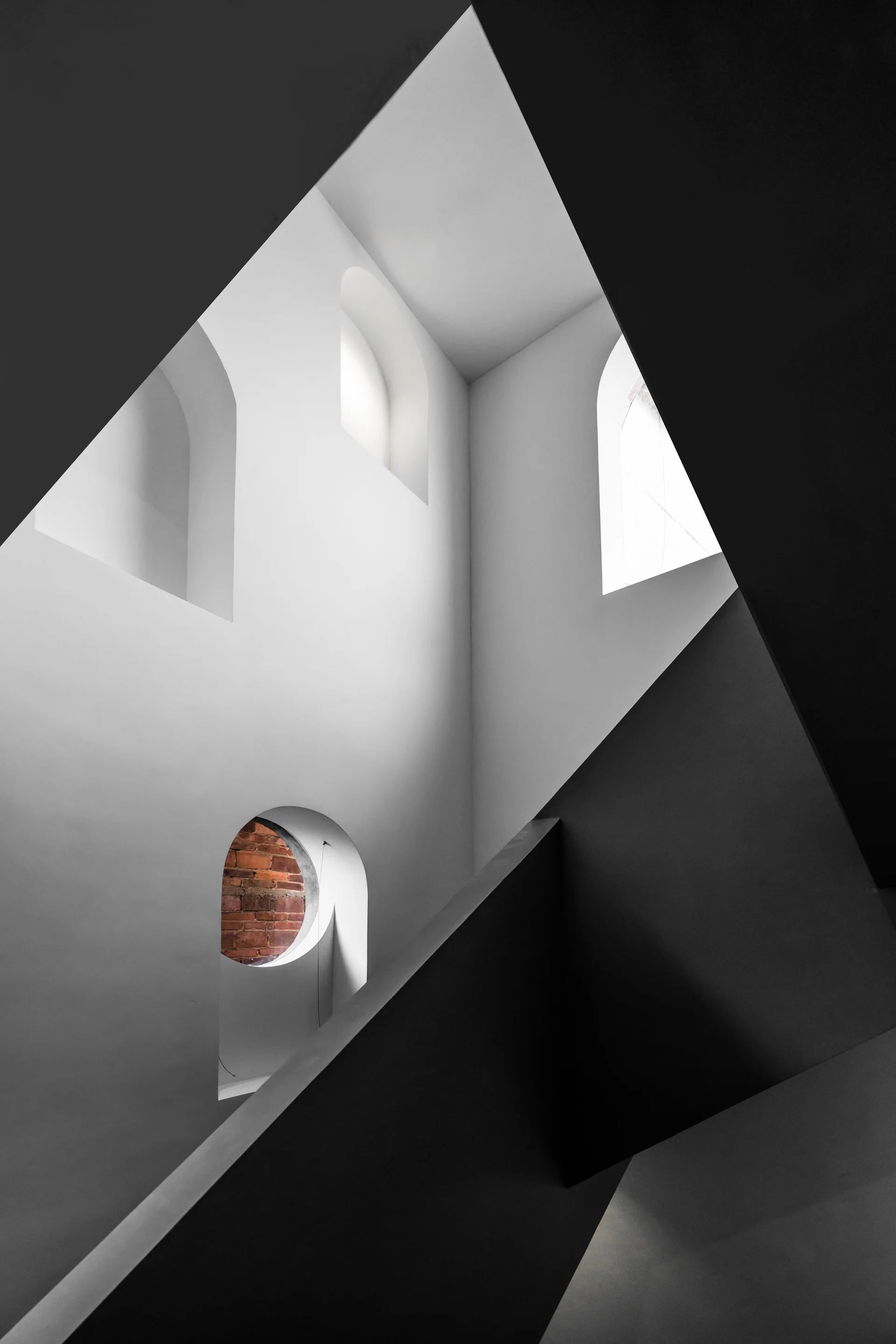 A minimalist white stairwell features sharp geometric shadow lines, arched window recesses, and a circular opening showing red brick.