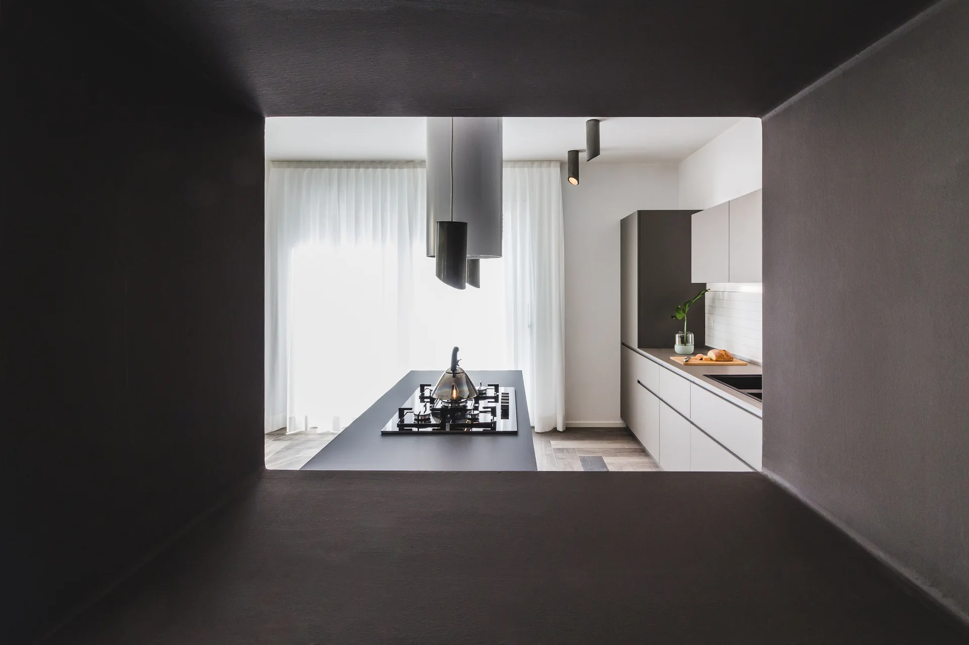A modern kitchen with a gray island and cabinetry is viewed through a dark, square wall opening under pendant lights.