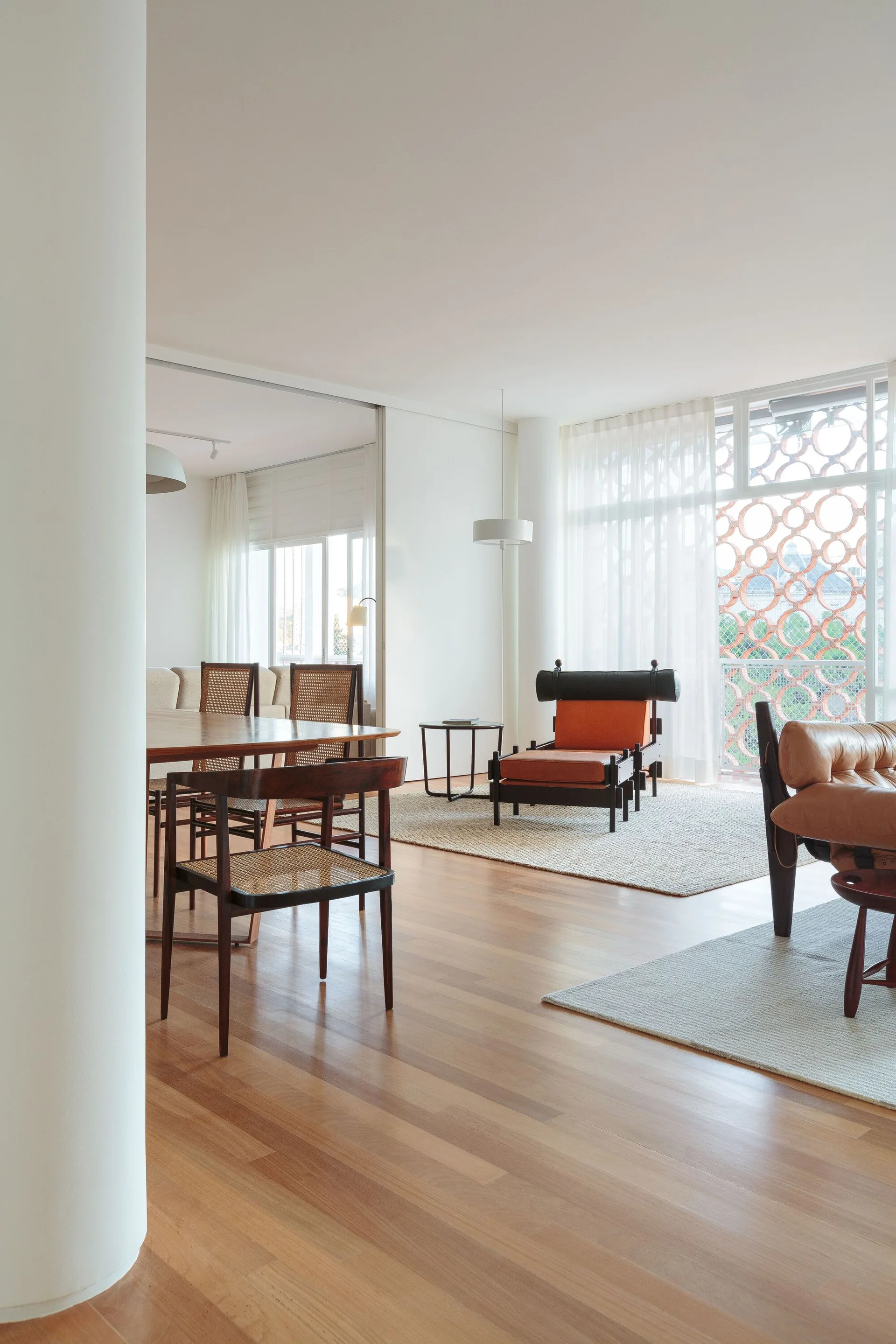 An open-plan dining space with a wooden table, cane chairs, wood flooring, and floor-to-ceiling windows filtered by sheer white curtains.
