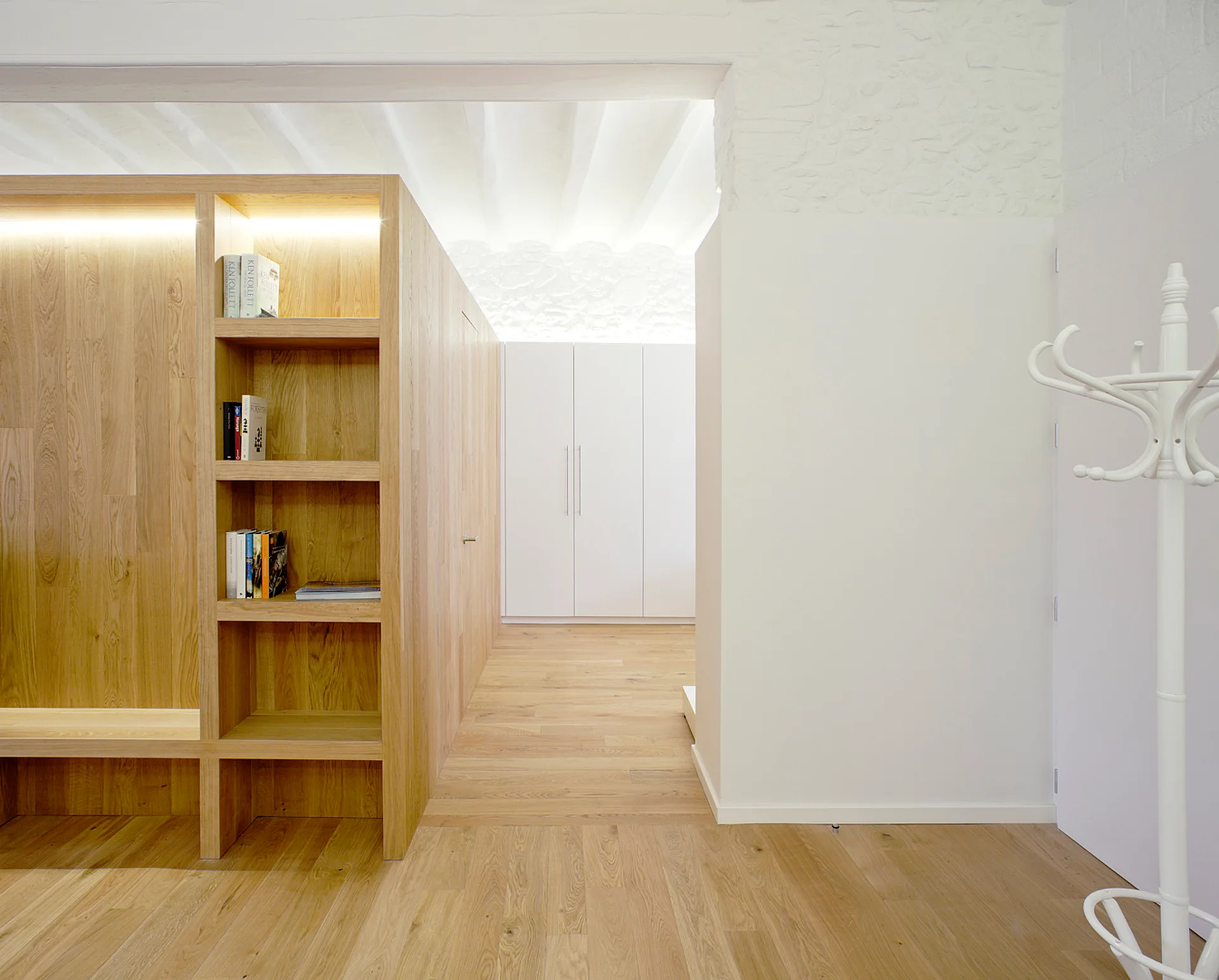 A modern minimalist hallway featuring a custom light wood shelving unit with integrated lighting, matching wood floors, and a white coat rack.