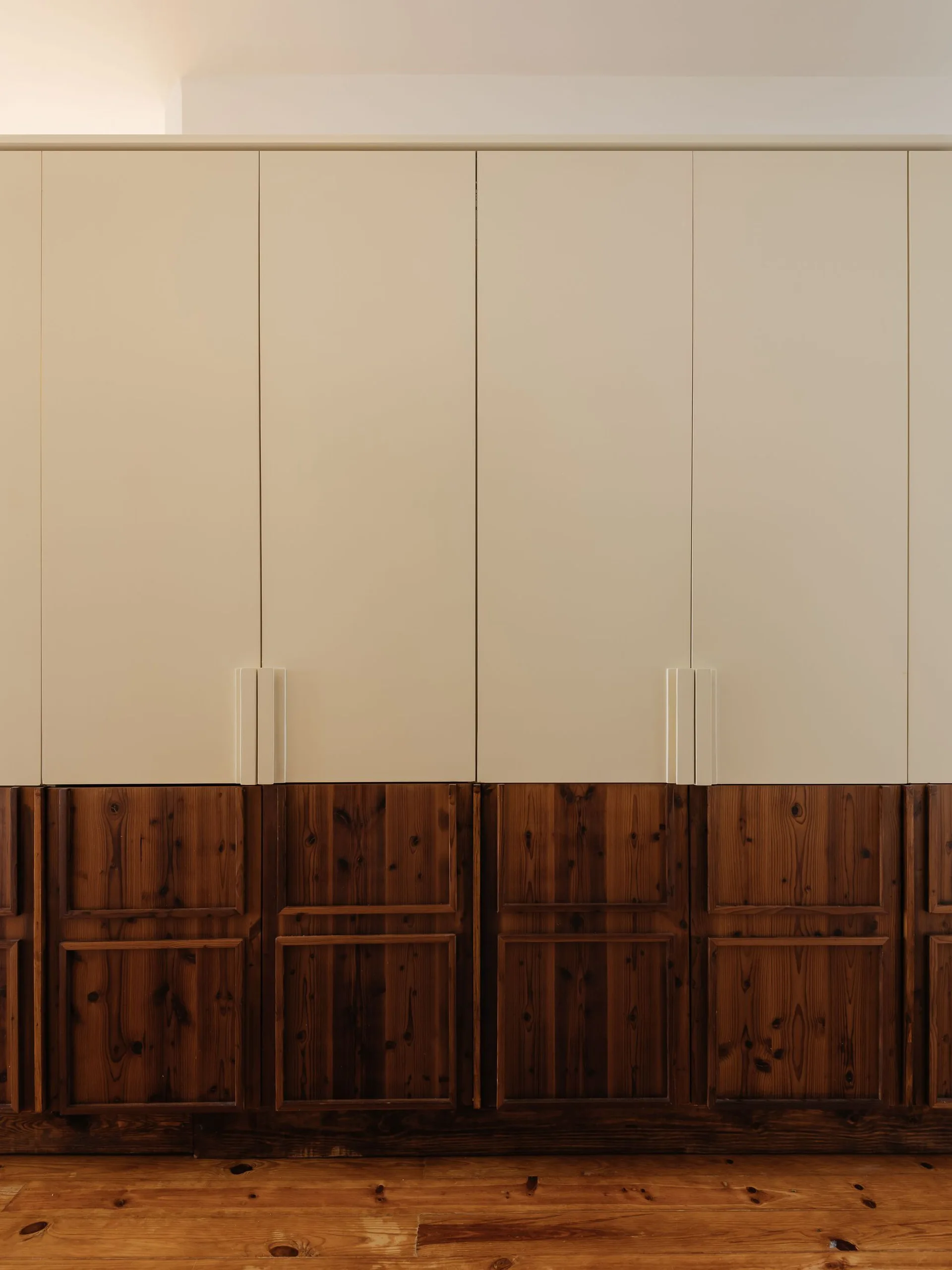 Floor-to-ceiling built-in wardrobe featuring smooth off-white upper doors with slim handles and knotty dark wood paneled lower doors over a wood floor.