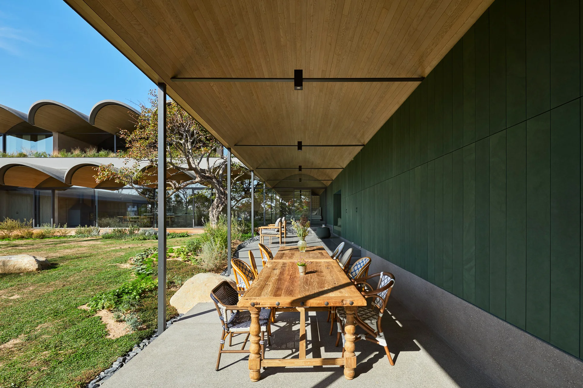 A long covered patio features a wooden dining table and chairs set against a deep green paneled wall beneath a wood plank ceiling.
