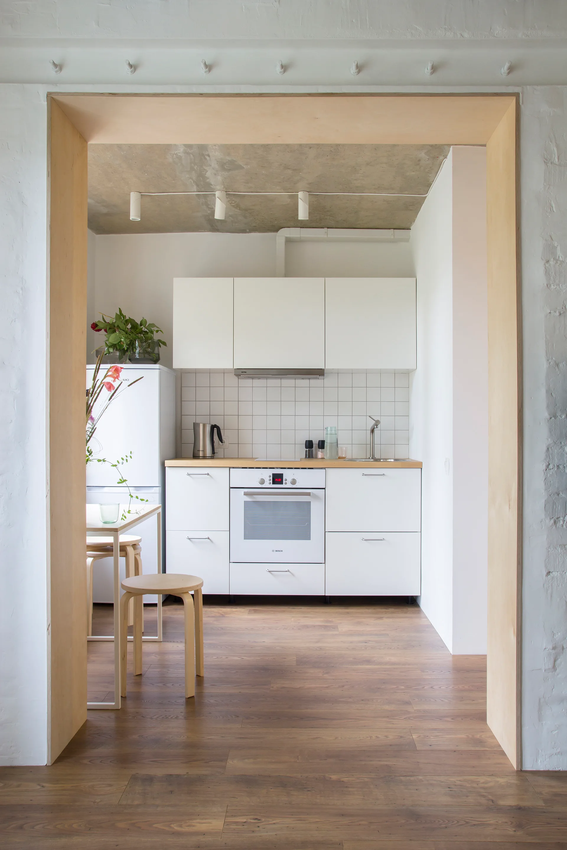 A view through a plywood portal into a minimalist white kitchen featuring flat-panel cabinets, wooden stools, and an exposed concrete ceiling.