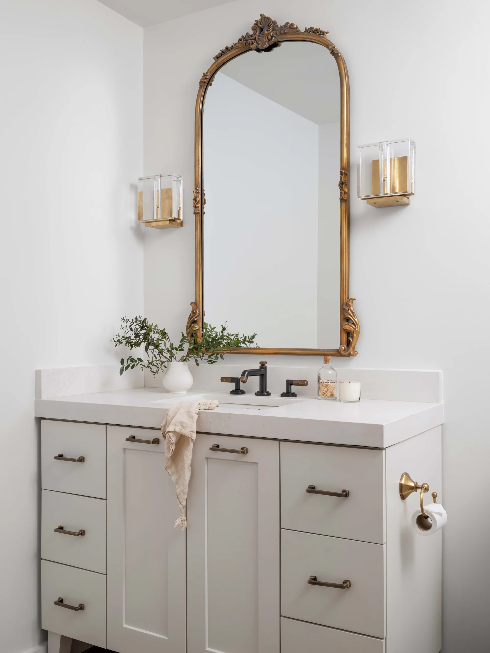 A white shaker-style bathroom vanity with a black faucet, topped by an arched ornate gold mirror and flanked by two glass sconces.