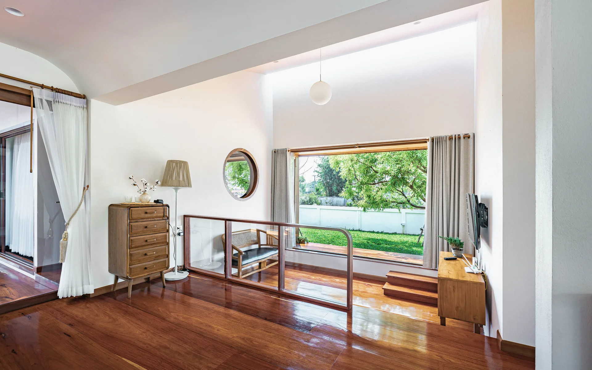 A split-level living room features polished hardwood flooring, a glass safety railing, and a large window overlooking a grassy yard.