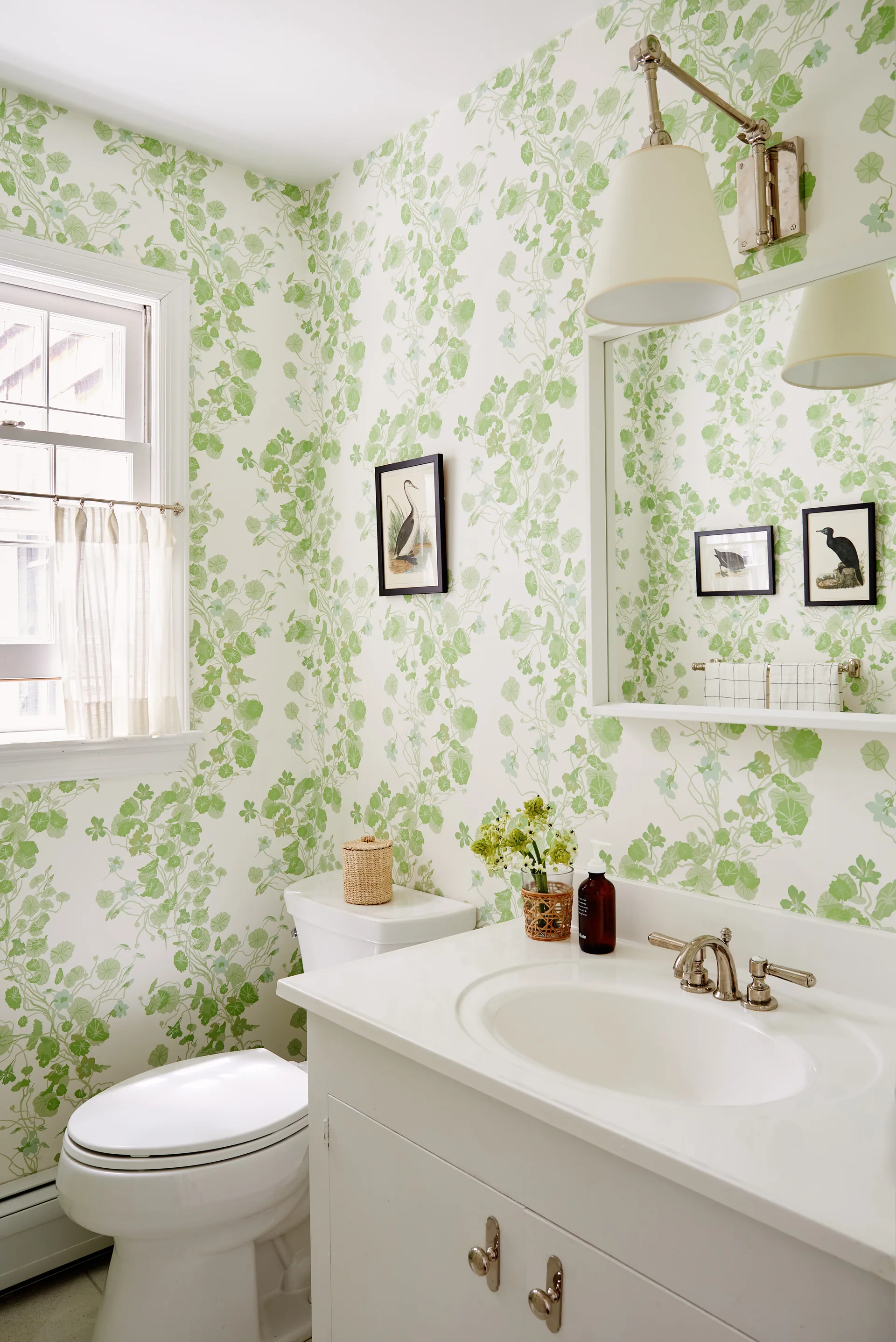 A bright powder room features green botanical wallpaper, a white vanity with silver fixtures, a matching toilet, and framed bird artwork.