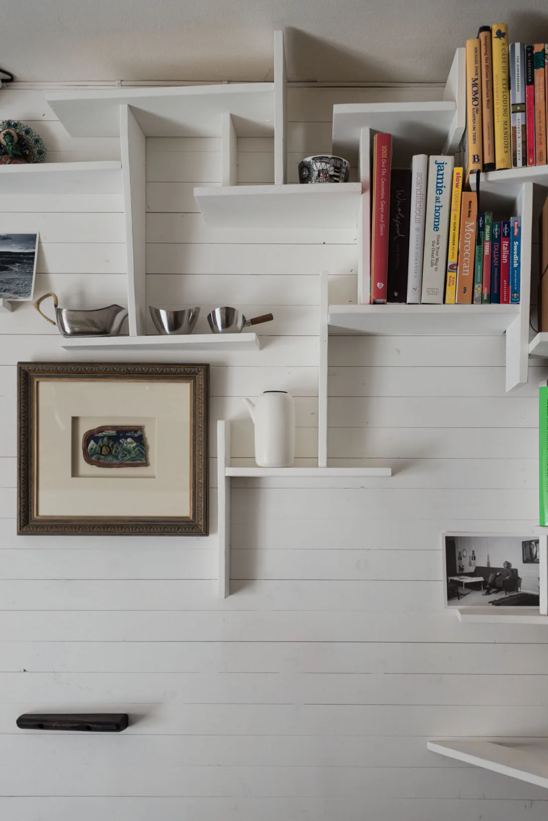 An asymmetric white wooden shelving unit is mounted on a white paneled wall and holds colorful books, small metal bowls, and framed art.
