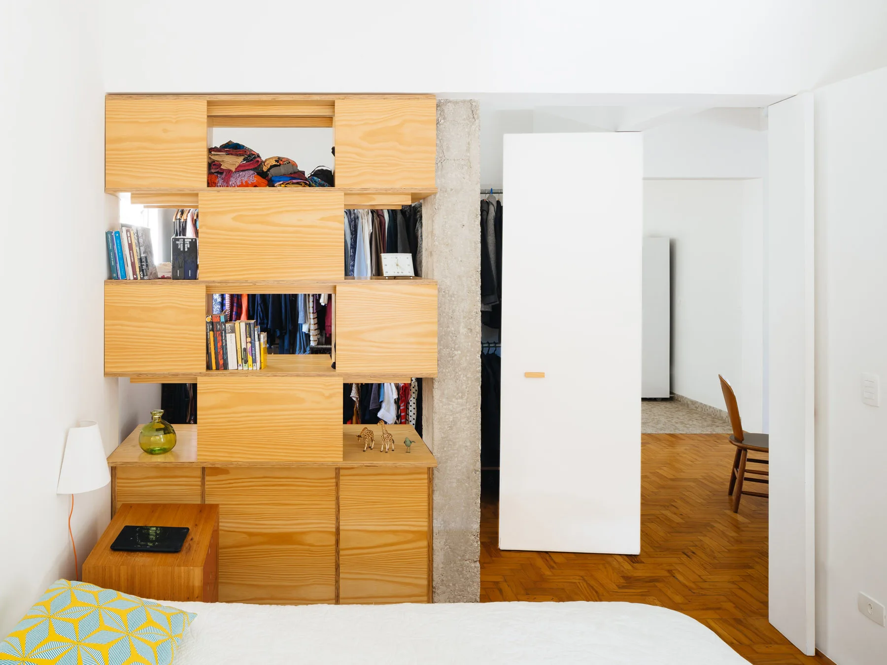 A bedroom features a light plywood modular shelving unit serving as a room divider next to an exposed concrete column and herringbone wood floors.