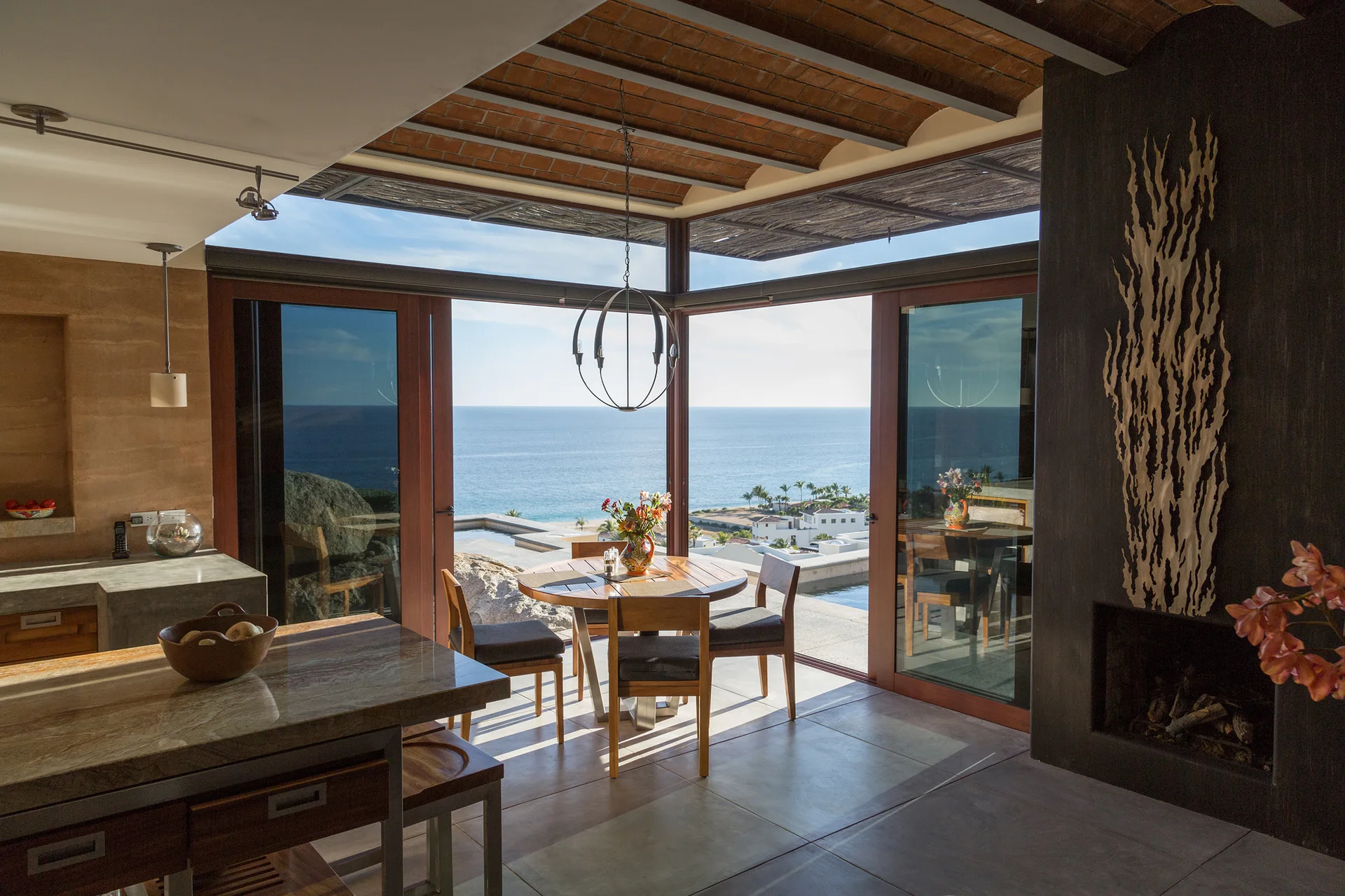 An indoor dining table and wooden chairs face sliding glass doors opening to a scenic ocean view beneath an exposed brick ceiling.