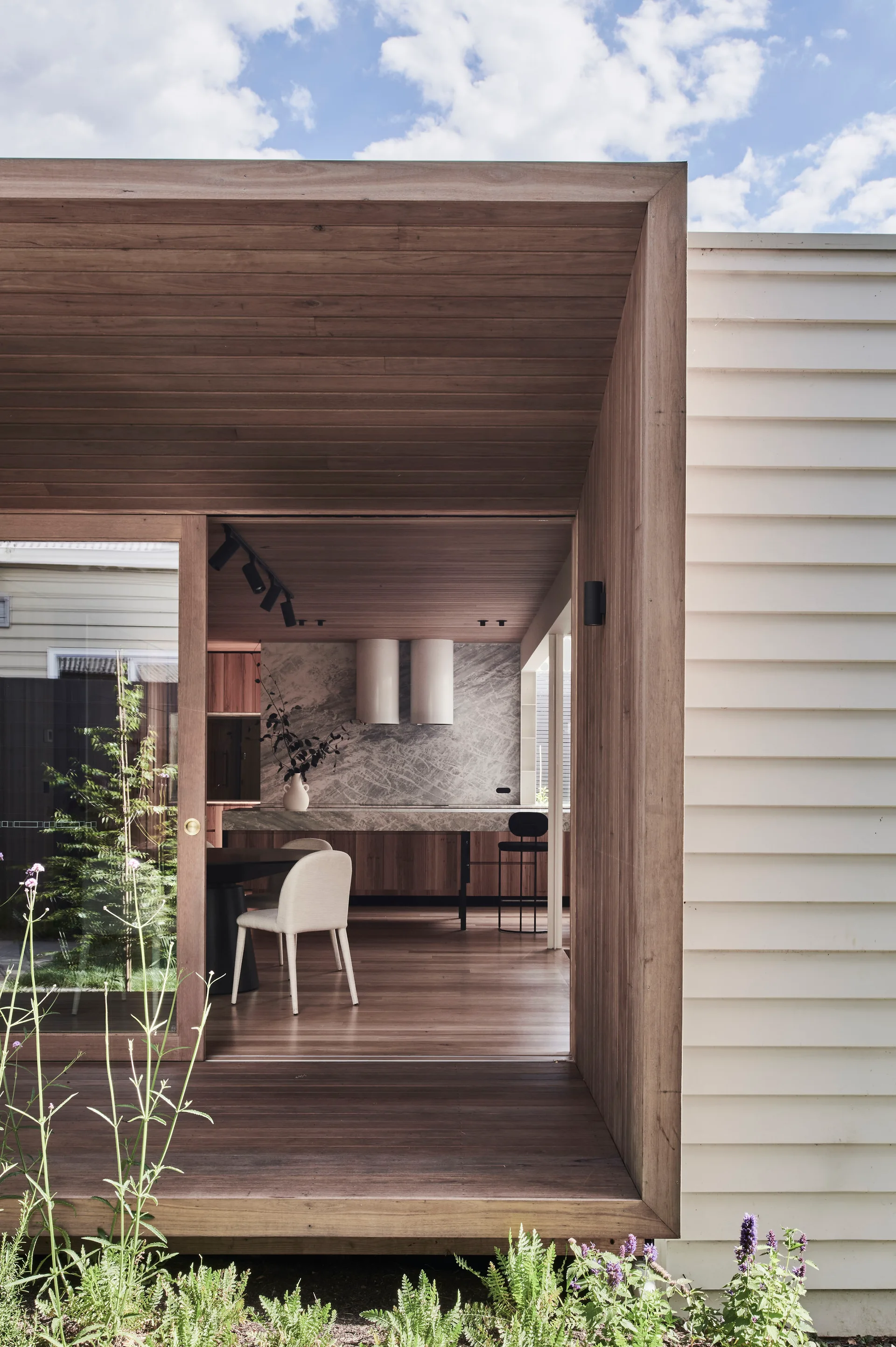 A view from a wooden deck through a large sliding door into a modern kitchen with wood walls, a marble island, and dining chairs.