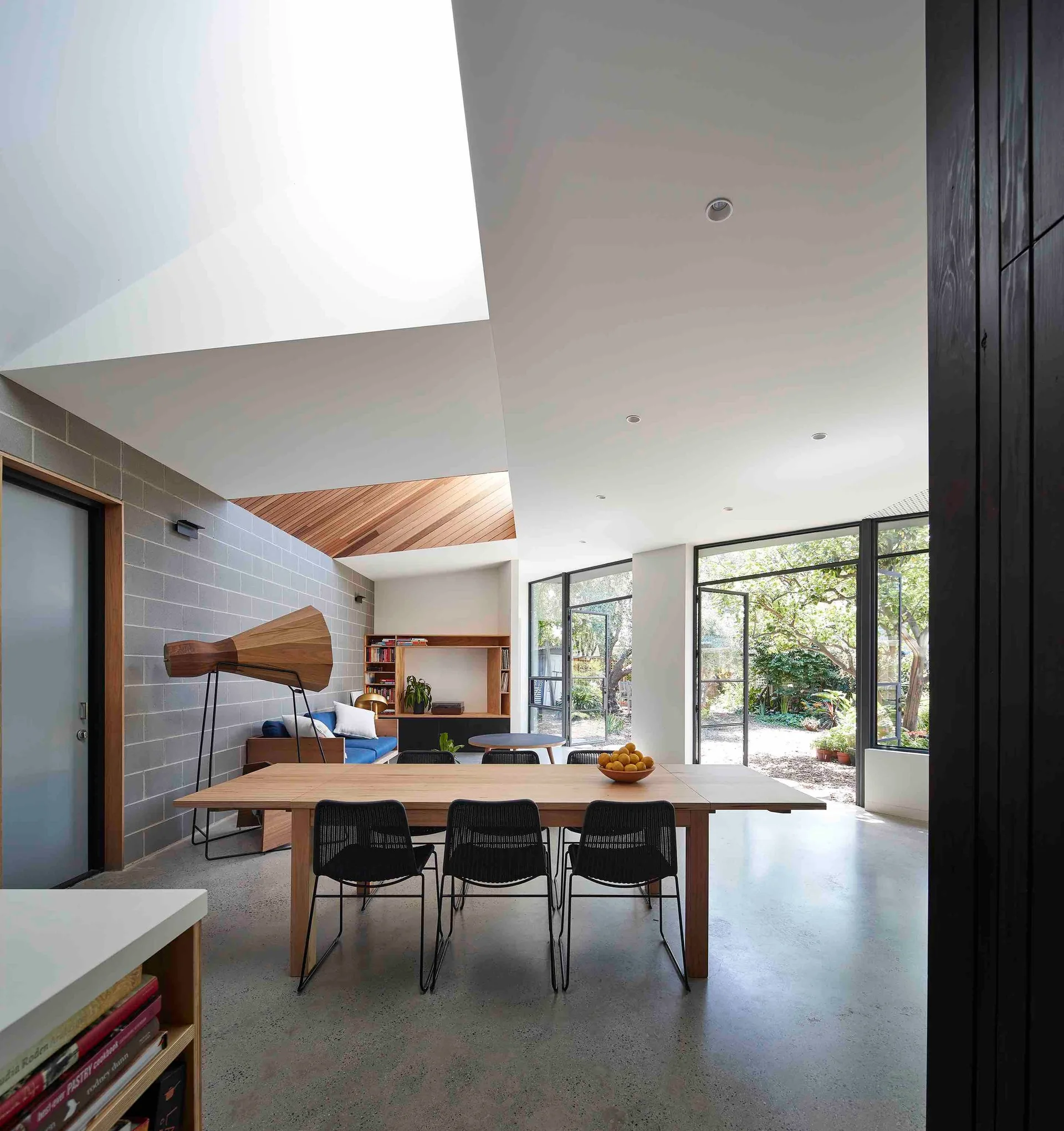 An open-plan dining room features a long wooden table with black chairs, a polished concrete floor, and an overhead angular skylight.