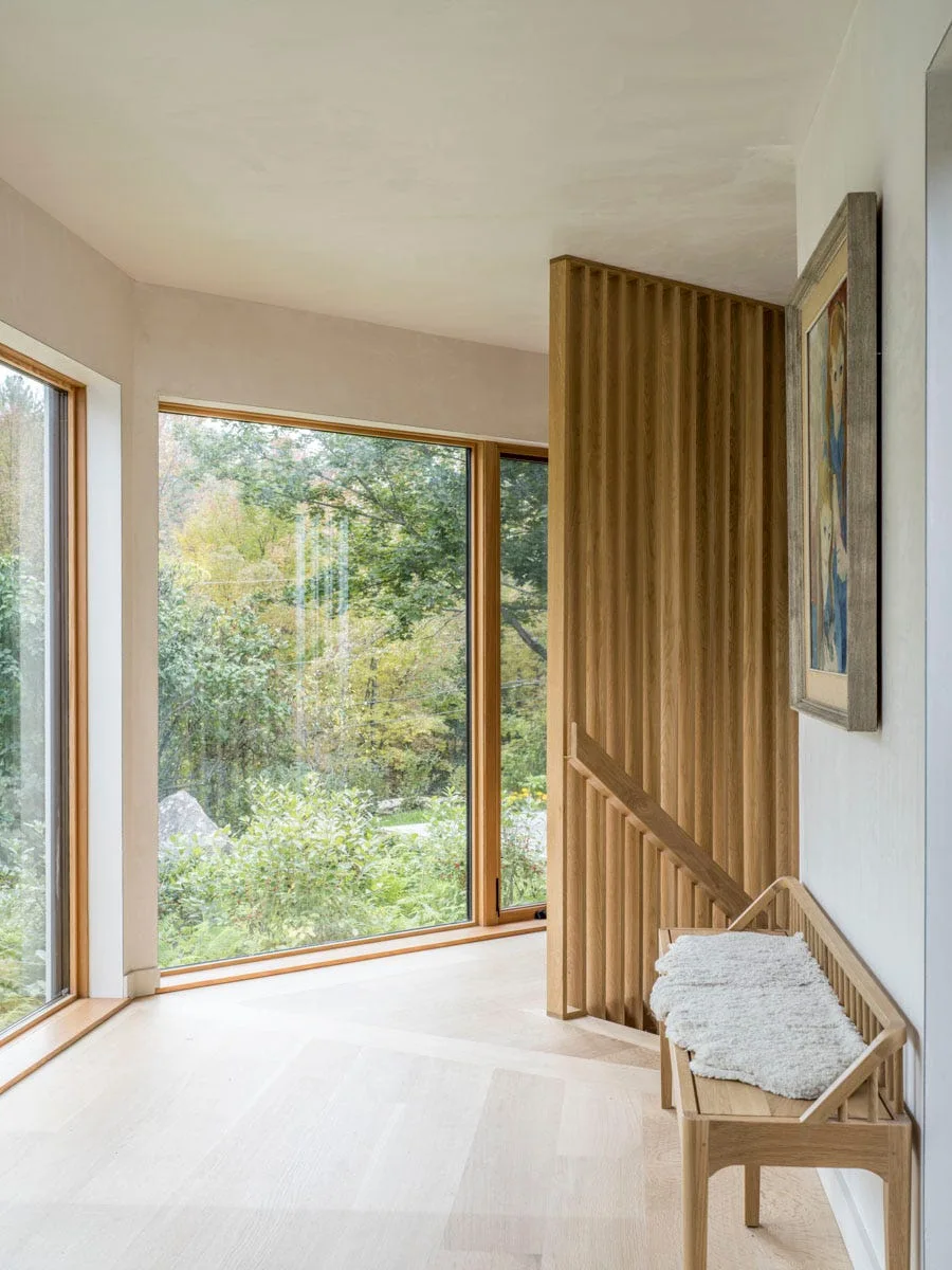 A bright entry area features a slatted light wood divider next to a wooden bench and large windows showing outdoor trees.
