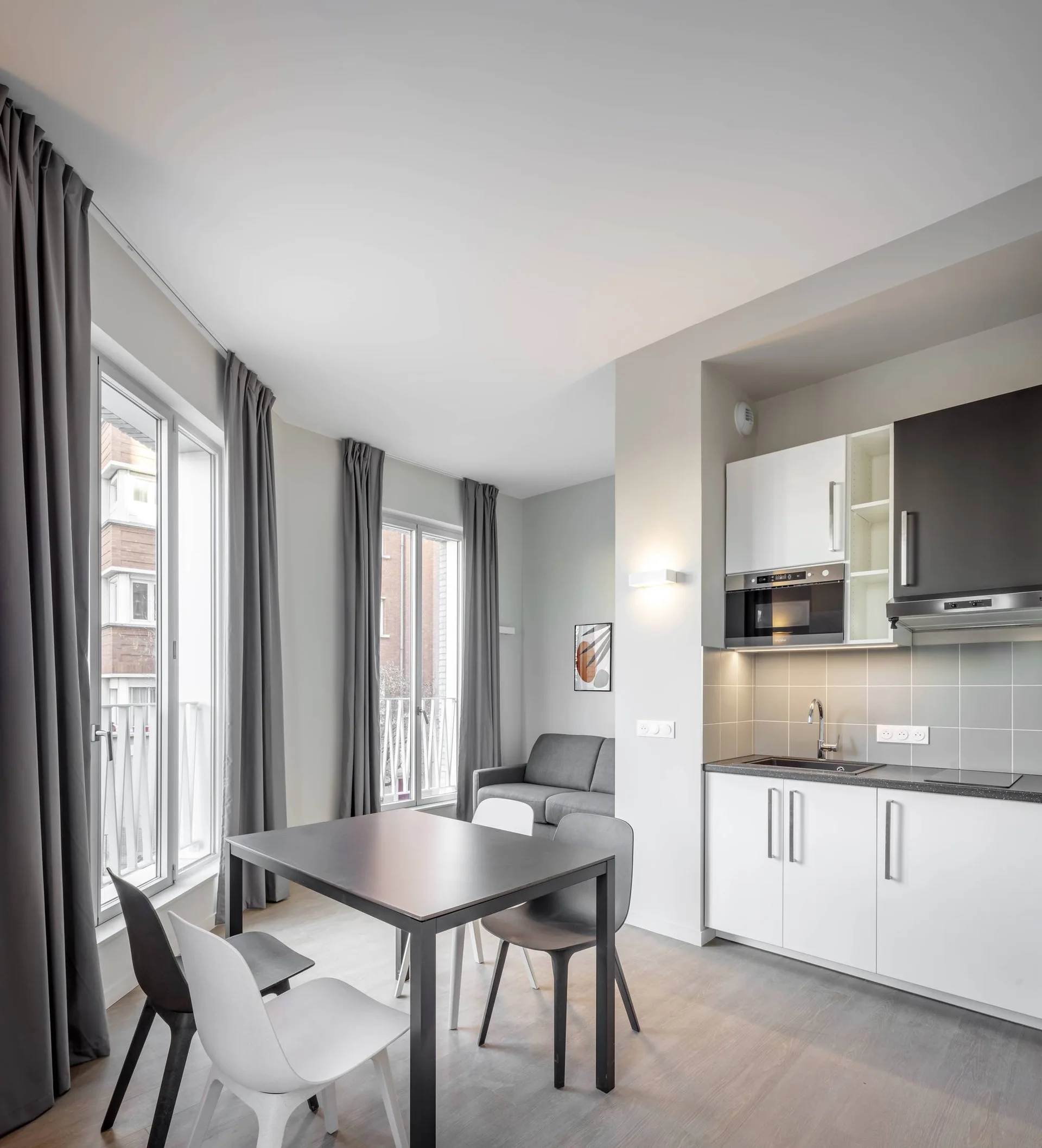 A modern studio apartment kitchen featuring white cabinetry, a grey tiled backsplash, a black dining table, and tall curtained windows.