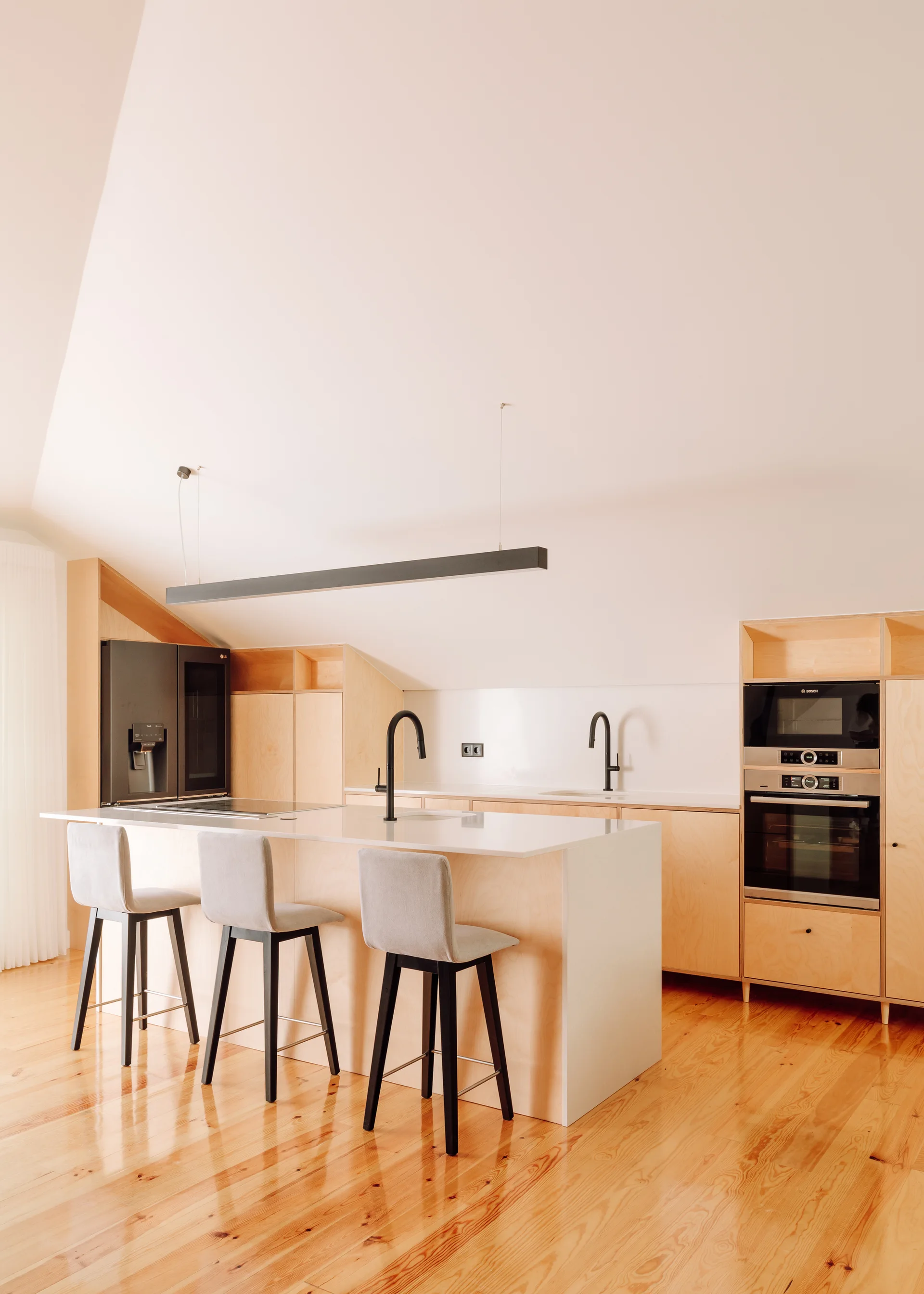 A modern kitchen with light plywood cabinets, a central island with three grey bar stools, black faucets, and a high sloped ceiling.
