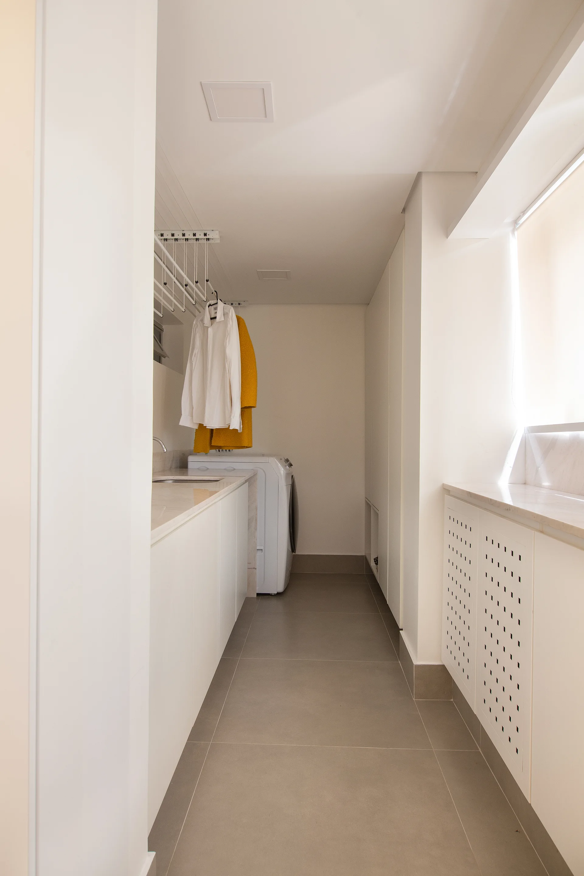 A narrow white laundry room features minimalist built-in cabinets, grey tile flooring, a utility sink, and a ceiling-mounted clothes drying rack.
