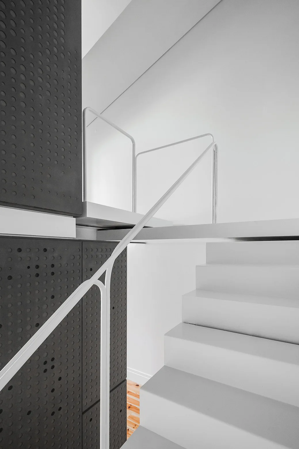 A modern white staircase with a minimalist thin metal railing stands next to dark grey perforated wall panels.