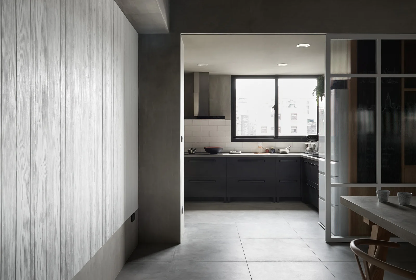 A modern kitchen featuring dark matte cabinets, white subway tile, a grey tiled floor, textured wall paneling, and a sliding fluted glass partition door.